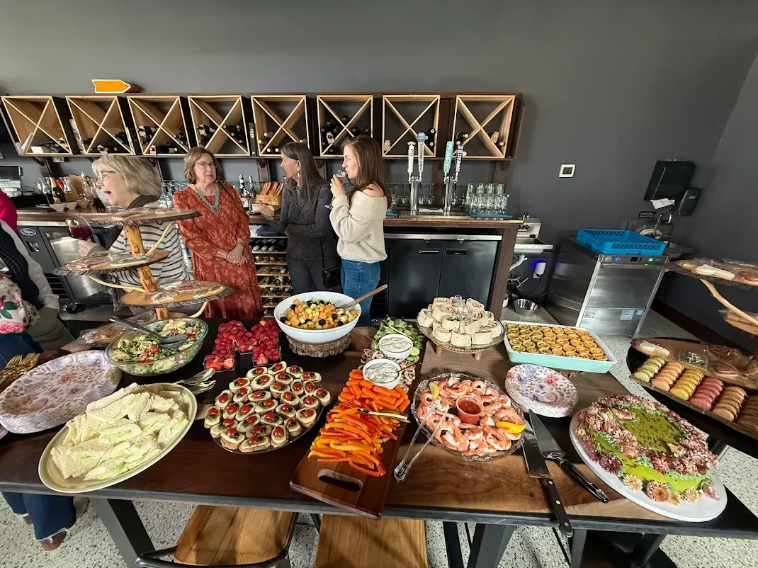 A buffet table with various salads, strawberries, layered sandwiches, sliced vegetables, shrimp, and a decorated cake at a gathering, with people talking in the background.