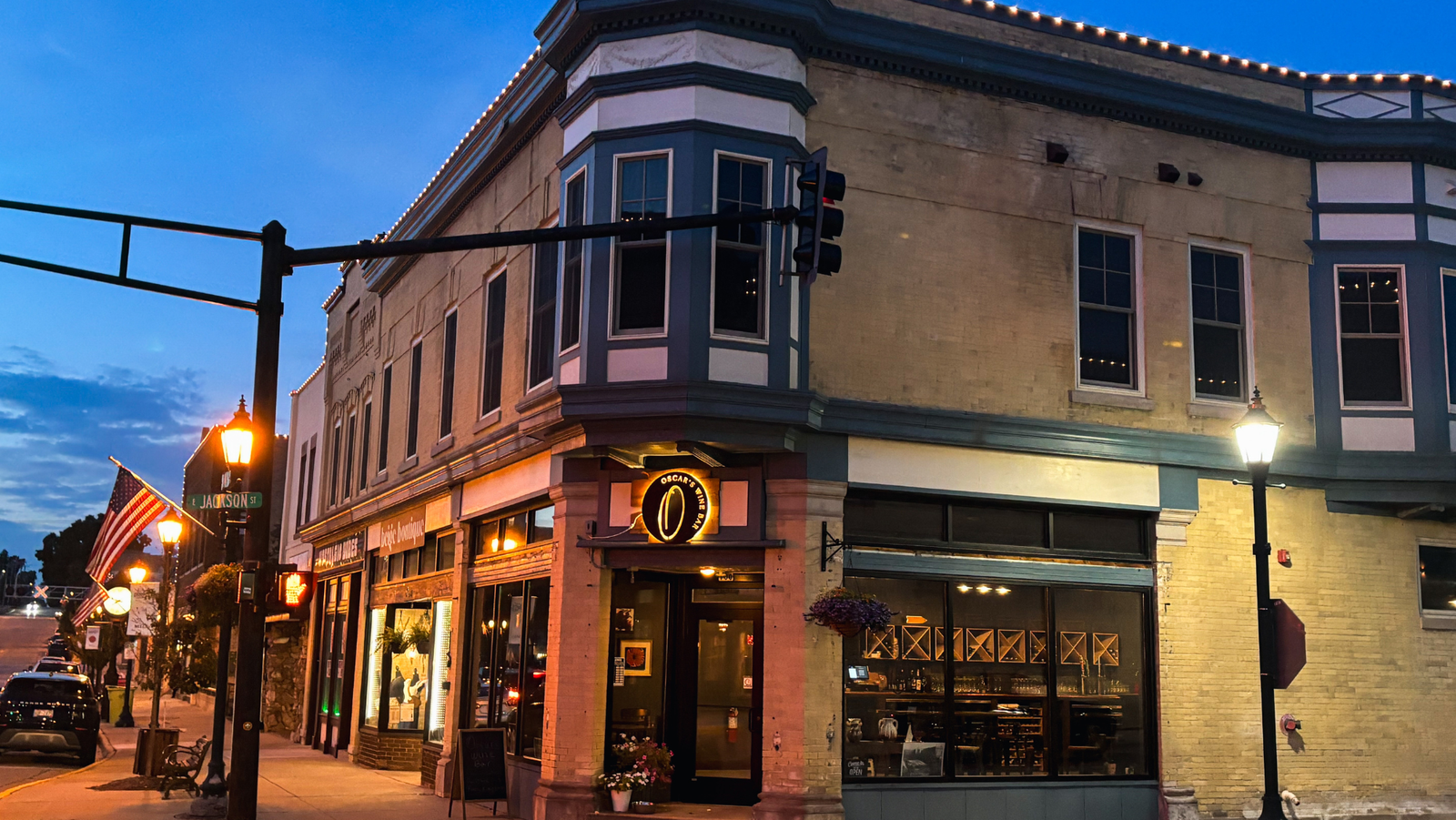 Corner building with restaurant named 'Oscar's' at night, American flags, street lamps, and traffic light, with illuminated windows and a sky transitioning from dusk to night.