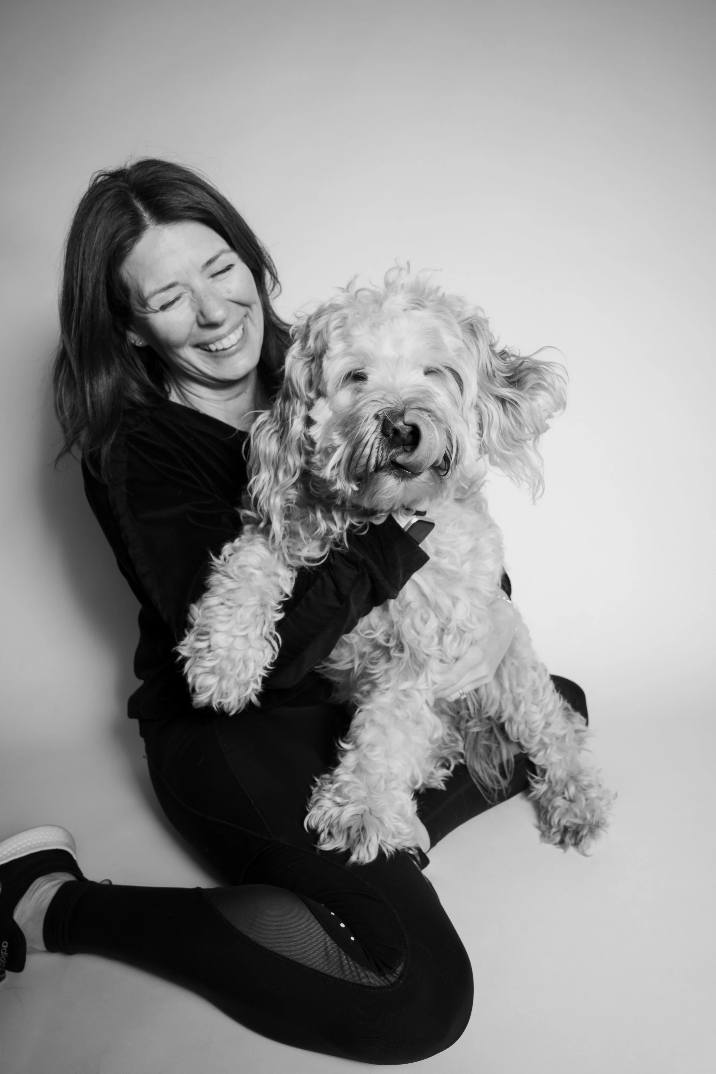 A woman with dark hair smiling while holding a large, fluffy dog with curly fur, against a plain light background.
