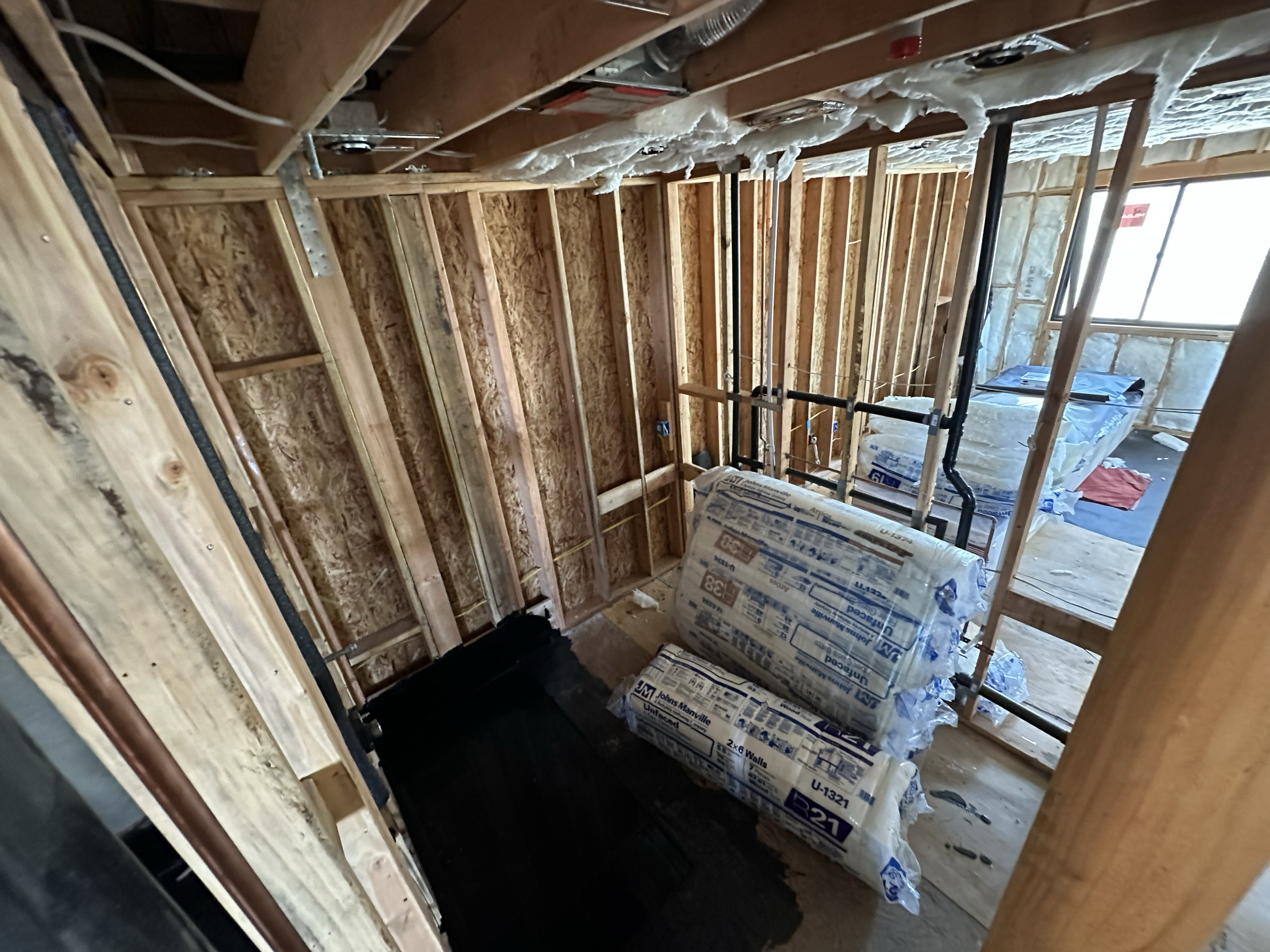 Interior view of a building under construction, showing exposed wooden framing, insulation, and construction materials. Window openings are visible, with some building supplies and tools present.