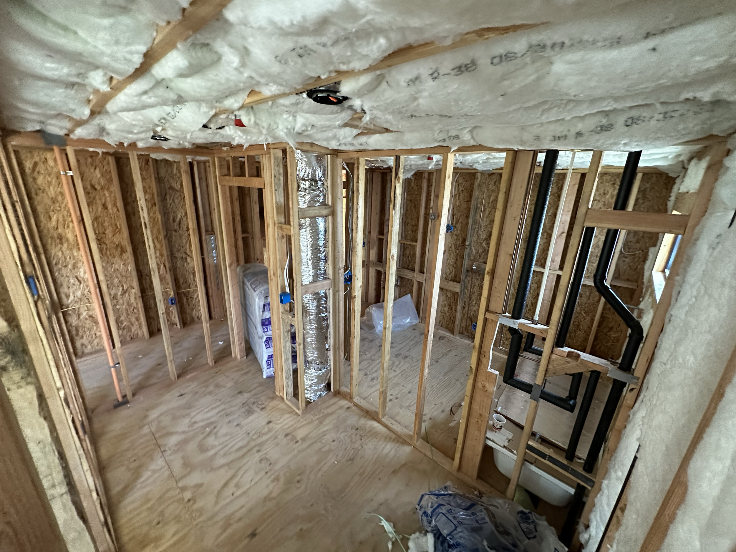 Interior view of a building under construction, showing exposed wooden framing, insulation installed on the ceiling, and visible heating or cooling ducts.
