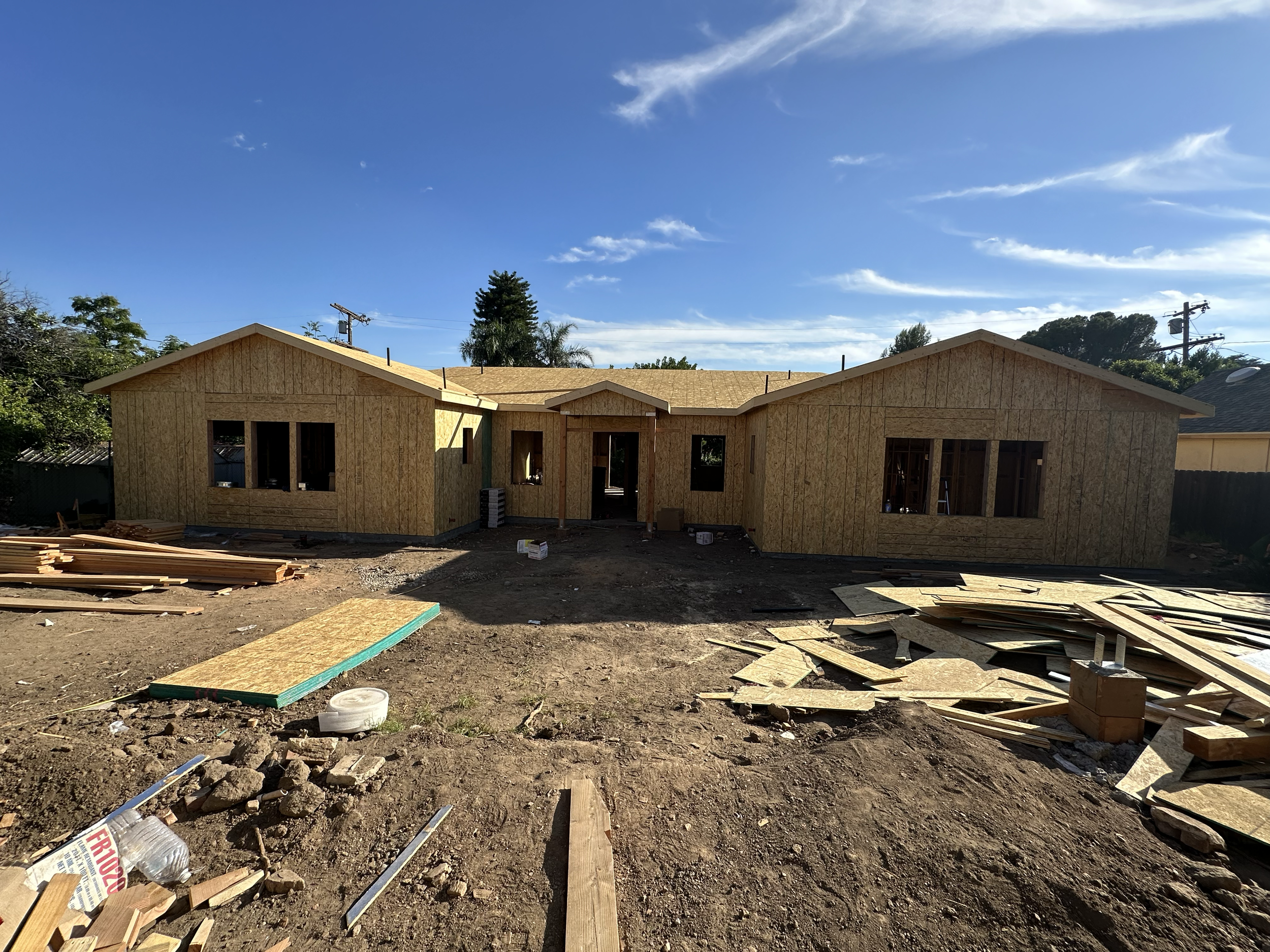 House under construction with wooden framework and scattered building materials and debris on the ground, under a partly cloudy blue sky.