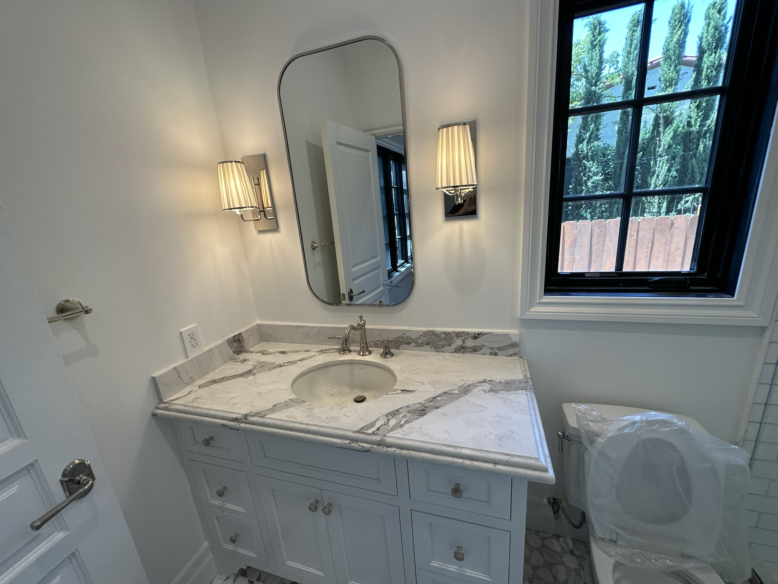 Bathroom with white cabinetry, marble countertop, oval sink, wall-mounted mirror, two wall sconces, window showing trees outside, and a toilet partially visible in the corner.