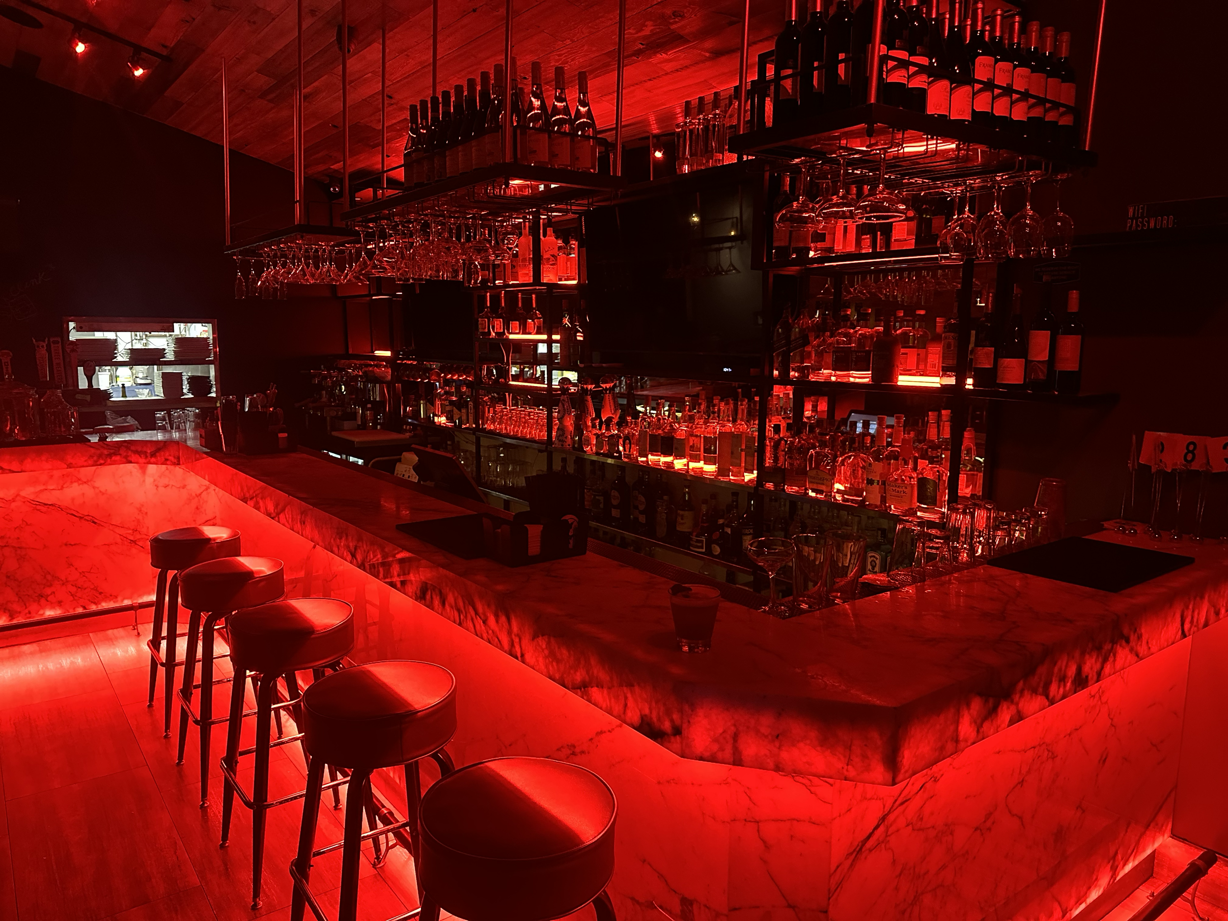 Bar area illuminated with red lighting, featuring wine bottles and glasses on racks, a marble counter with bar stools, and a darkened background.