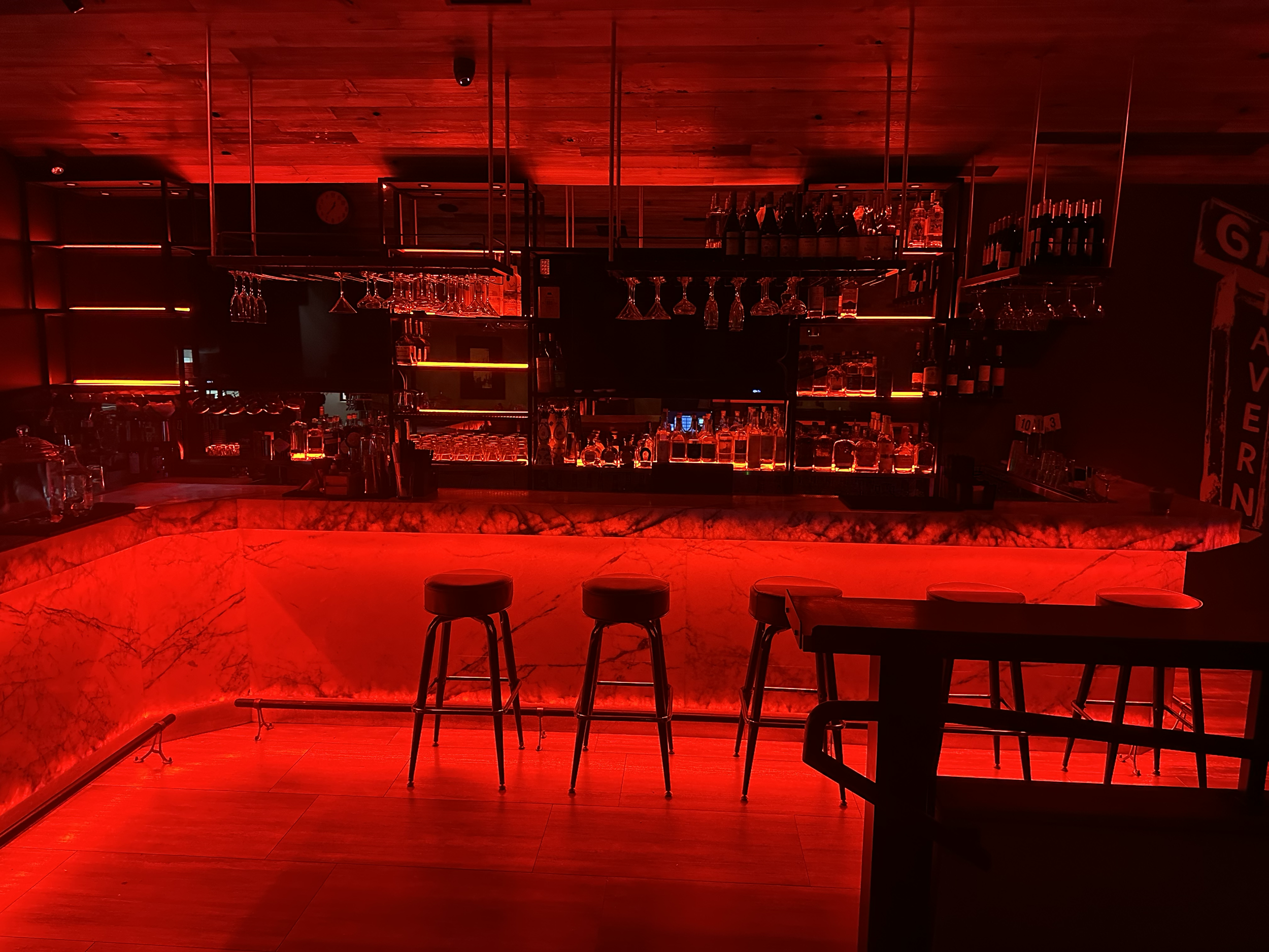A bar area illuminated with red lighting, featuring a marble countertop, bar stools, glassware hanging upside down, and shelves with bottles of alcohol.