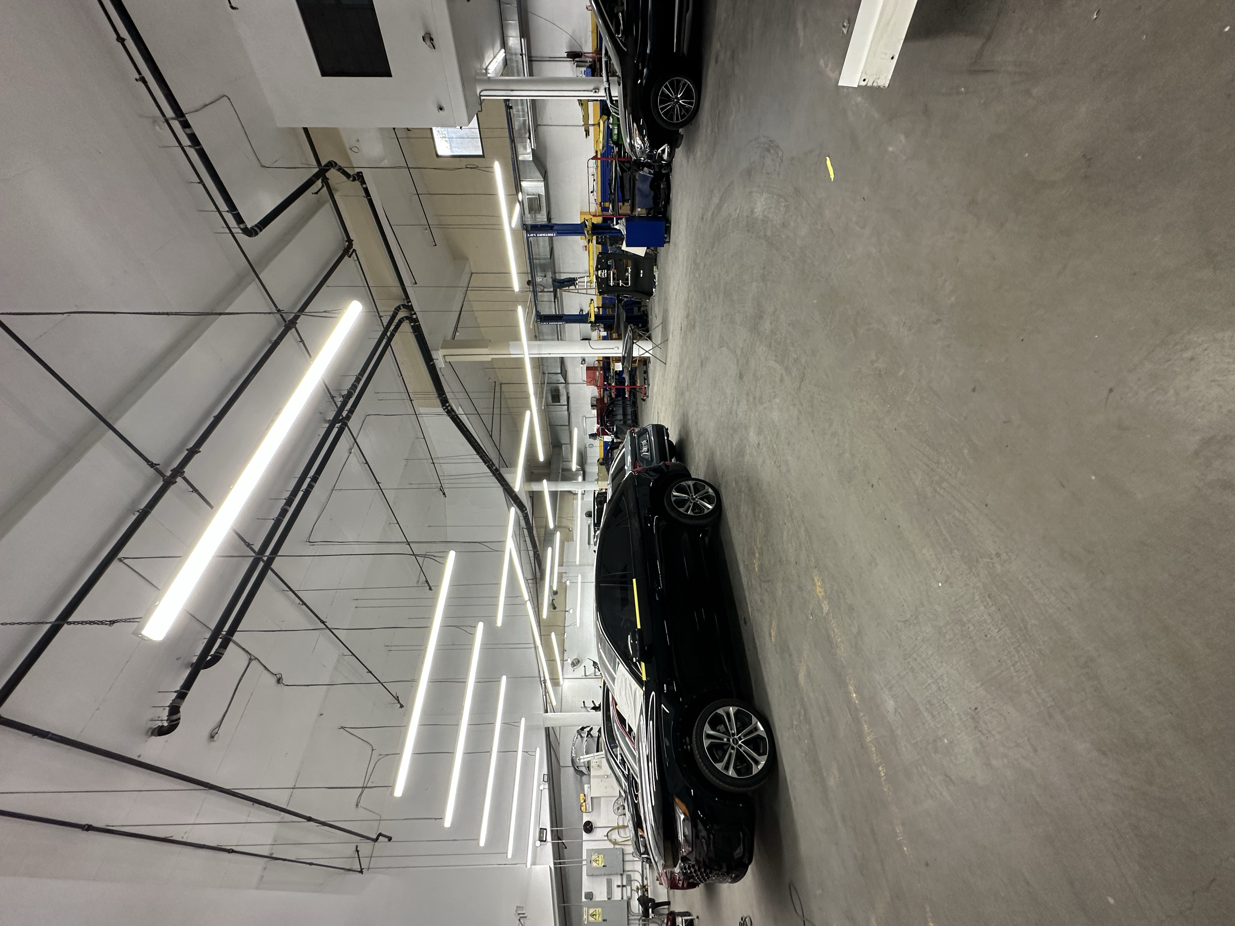 An indoor car wash or detailing facility with black cars parked and equipment around, including bright lighting and pipes on the ceiling.