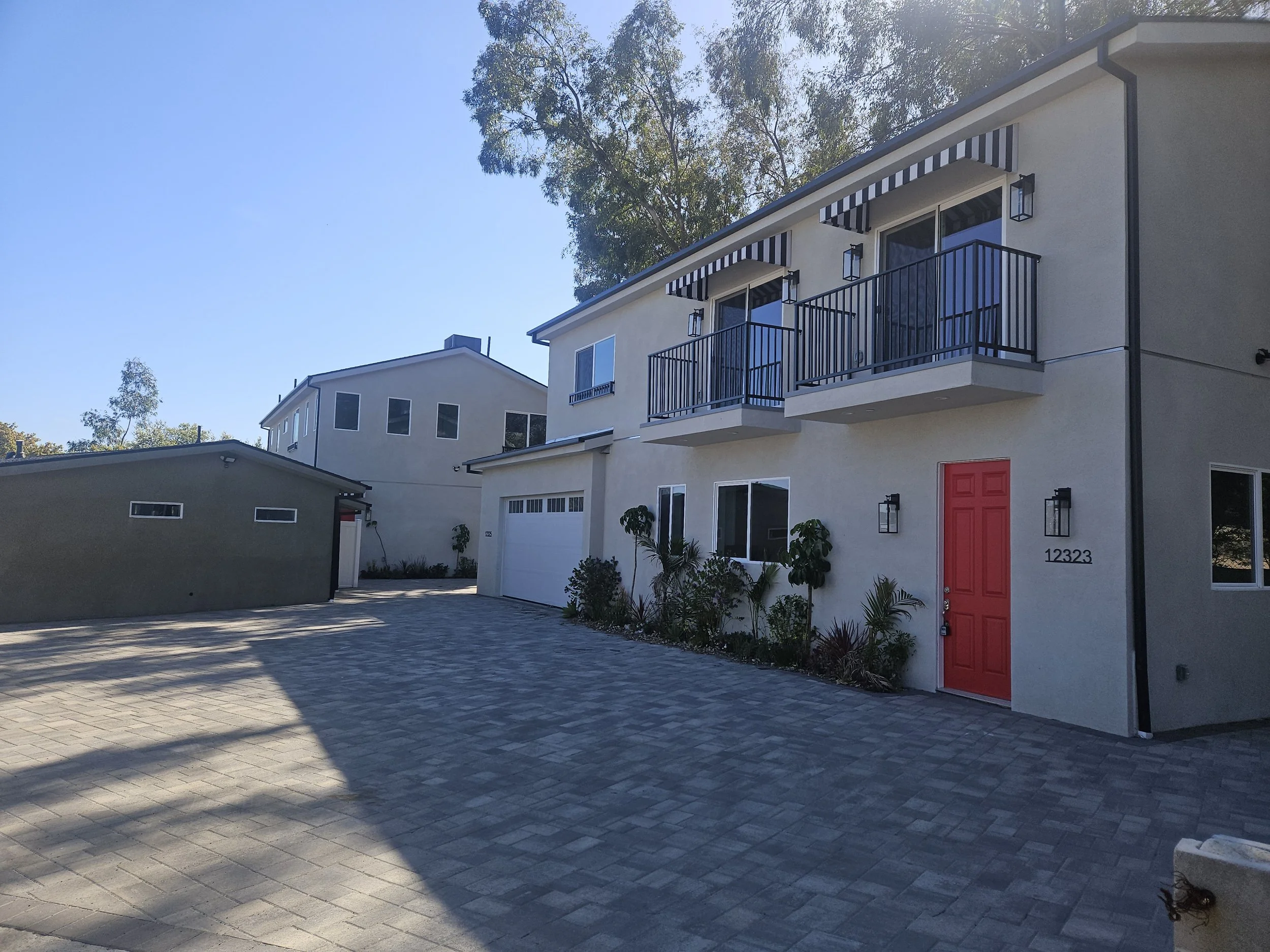 A modern residential building with white stucco walls, red front door, black metal balconies, and striped window awnings. The house has a driveway with gray pavers and outdoor lighting fixtures.