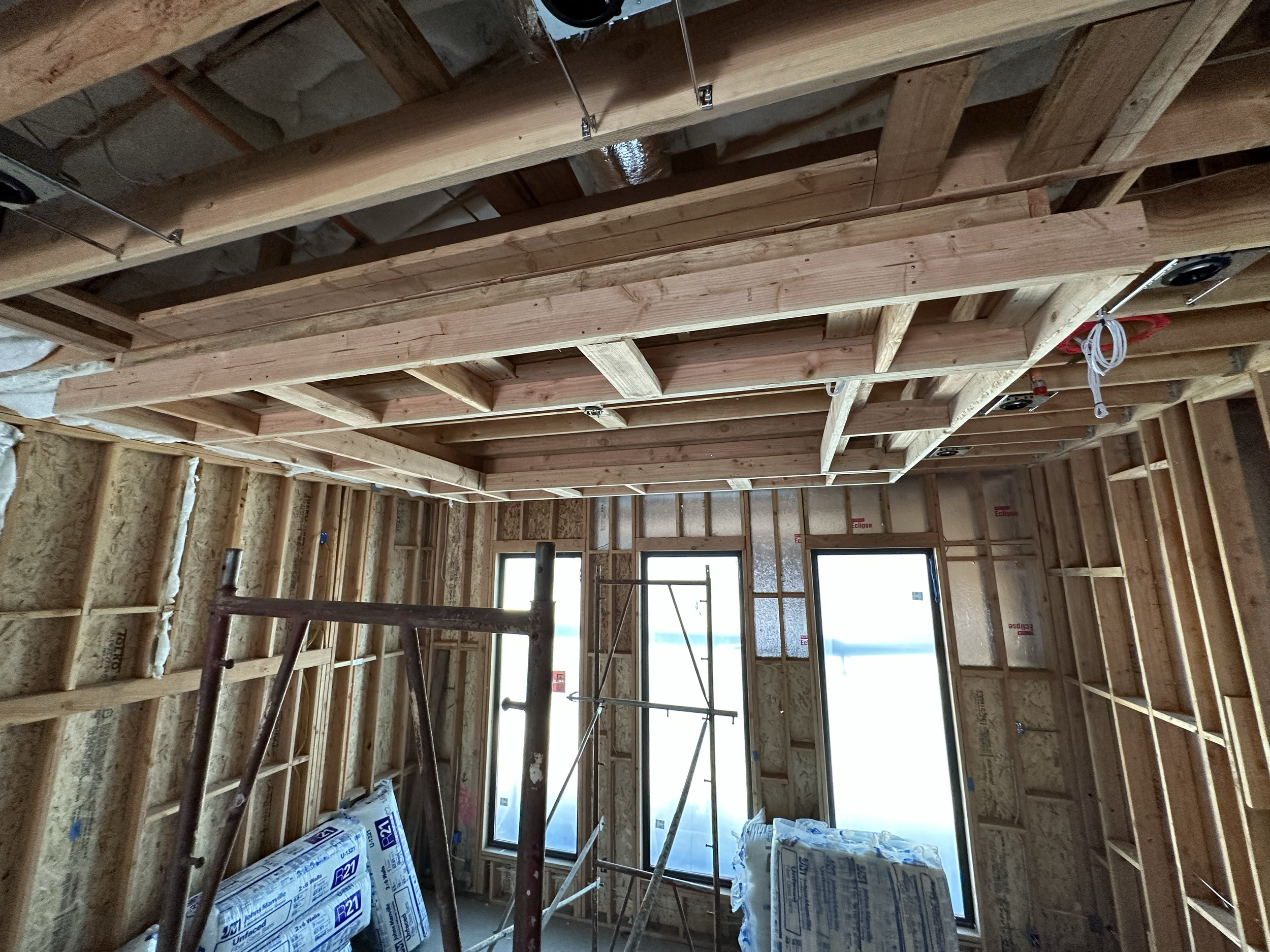 Interior view of a building under construction with exposed wooden framing, insulation, and windows.