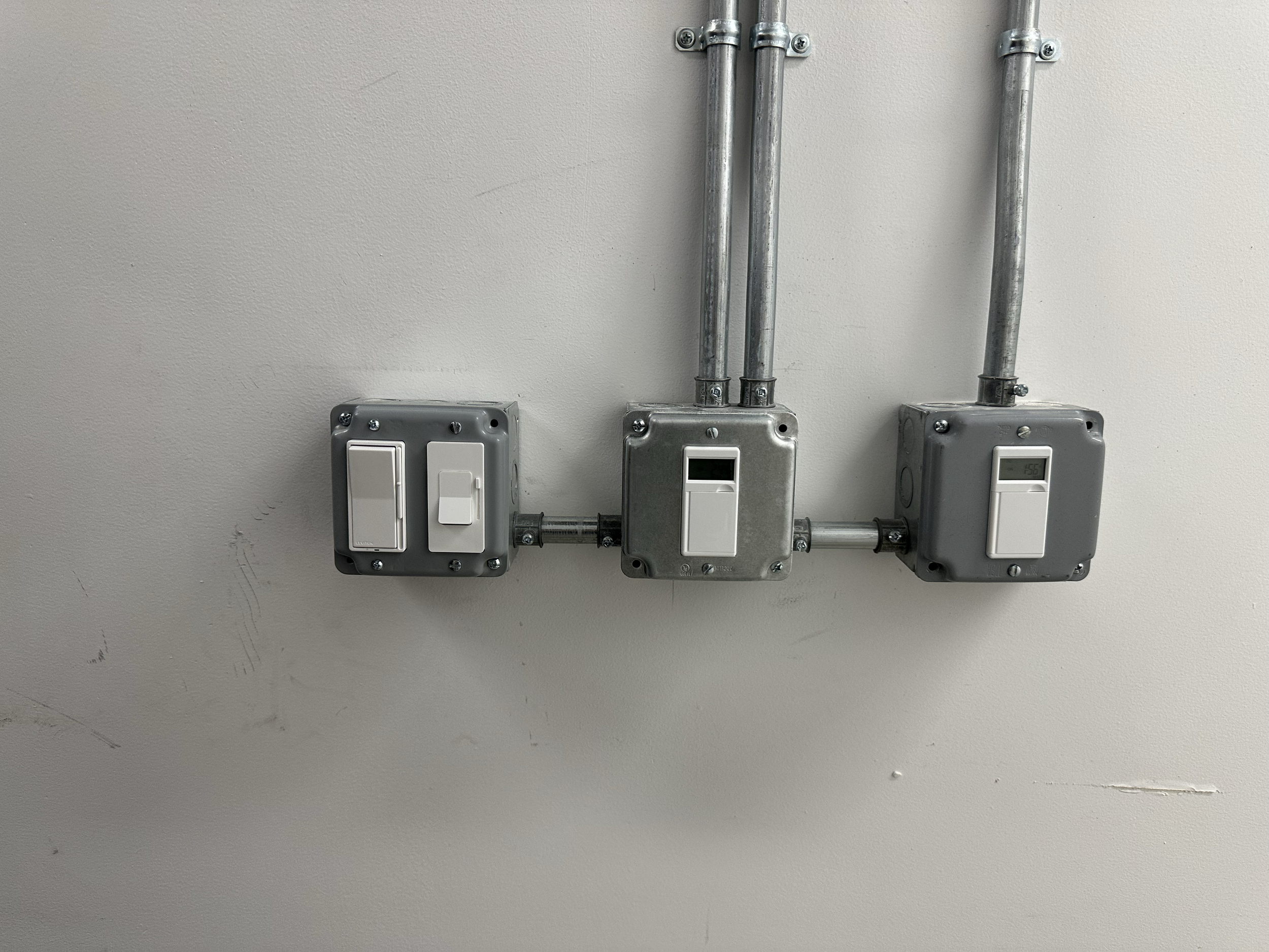 Three electrical switchboxes with digital meters and electrical conduit pipes mounted on a beige wall.