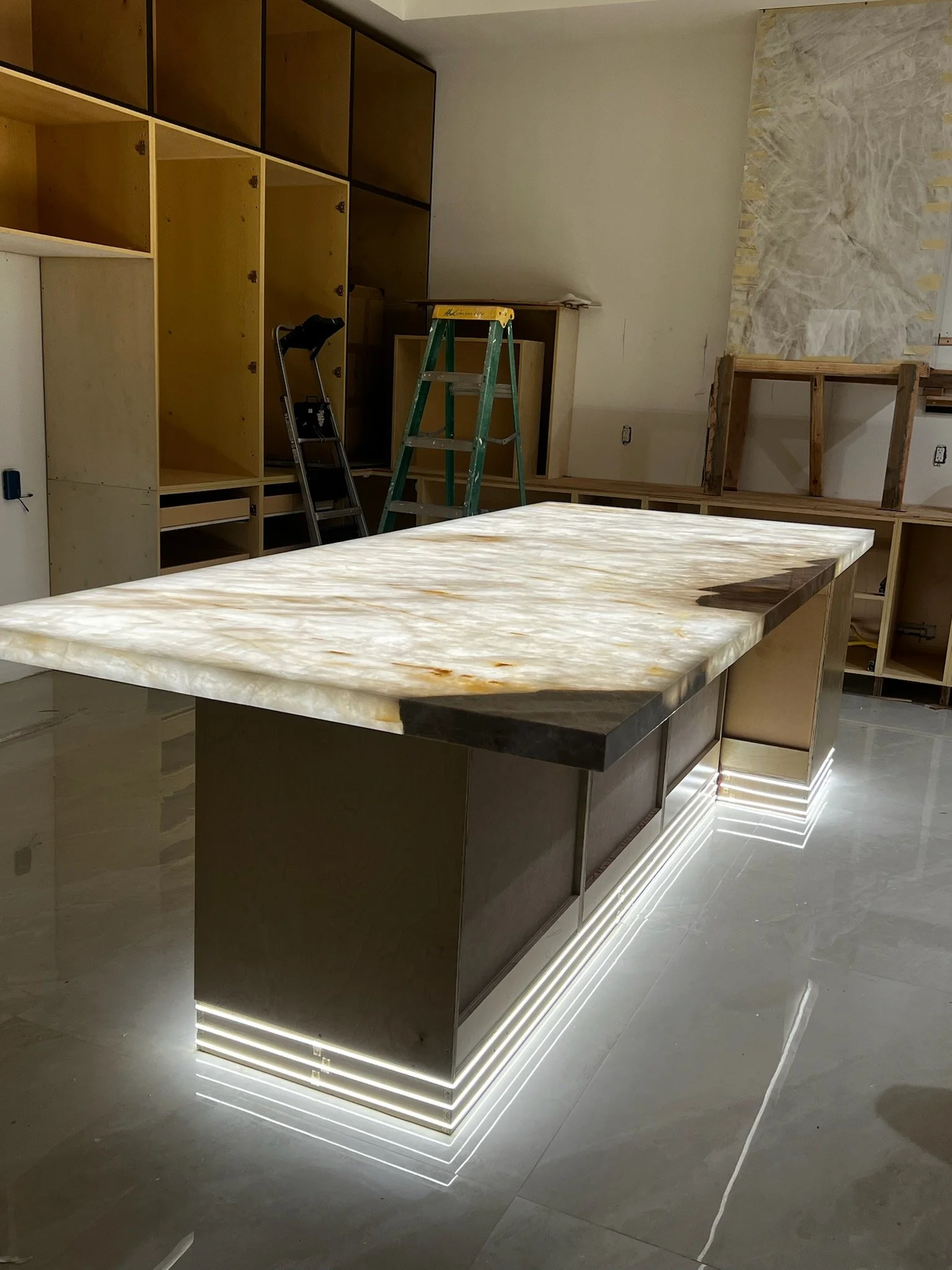 Interior of a room under construction with an island featuring a white marble countertop, surrounded by open shelving and cabinetry, with a ladder and tools visible.