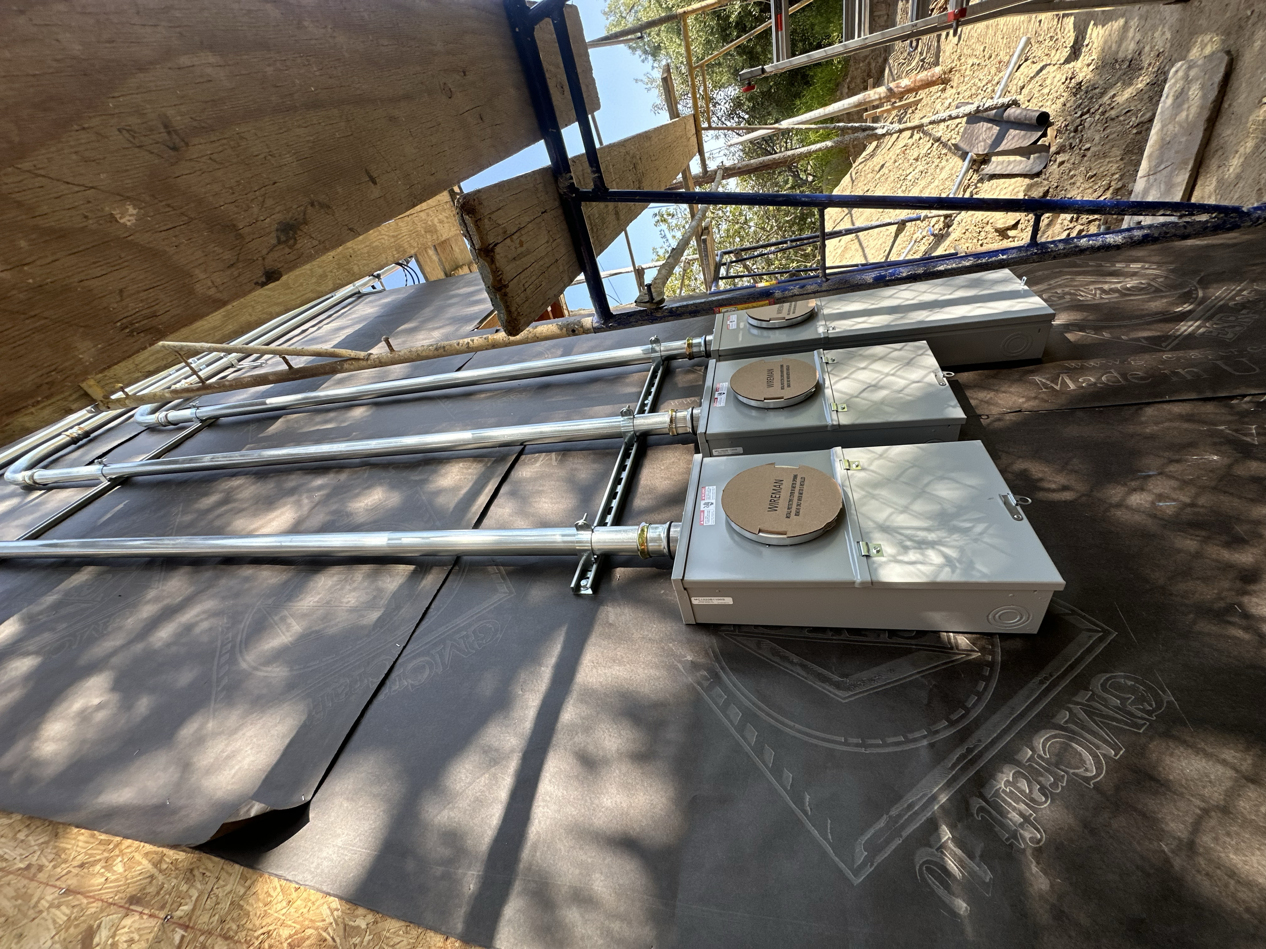 Construction site with electrical meters, metal pipes, wooden framework, and scaffolding outdoors with trees and blue sky in the background.