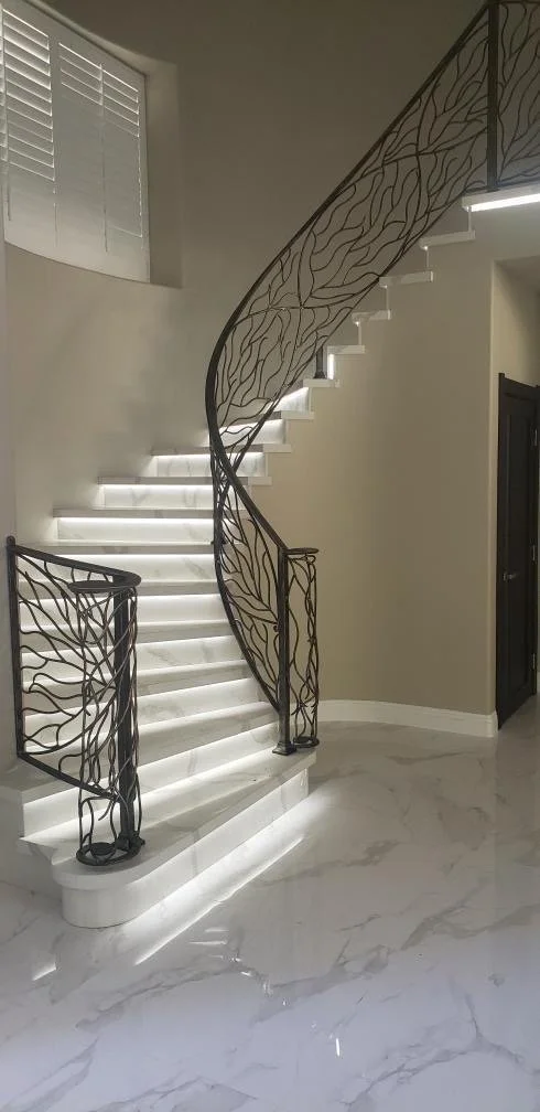 Elegant white spiral staircase with illuminated steps and decorative black wrought iron railing in a modern home interior.
