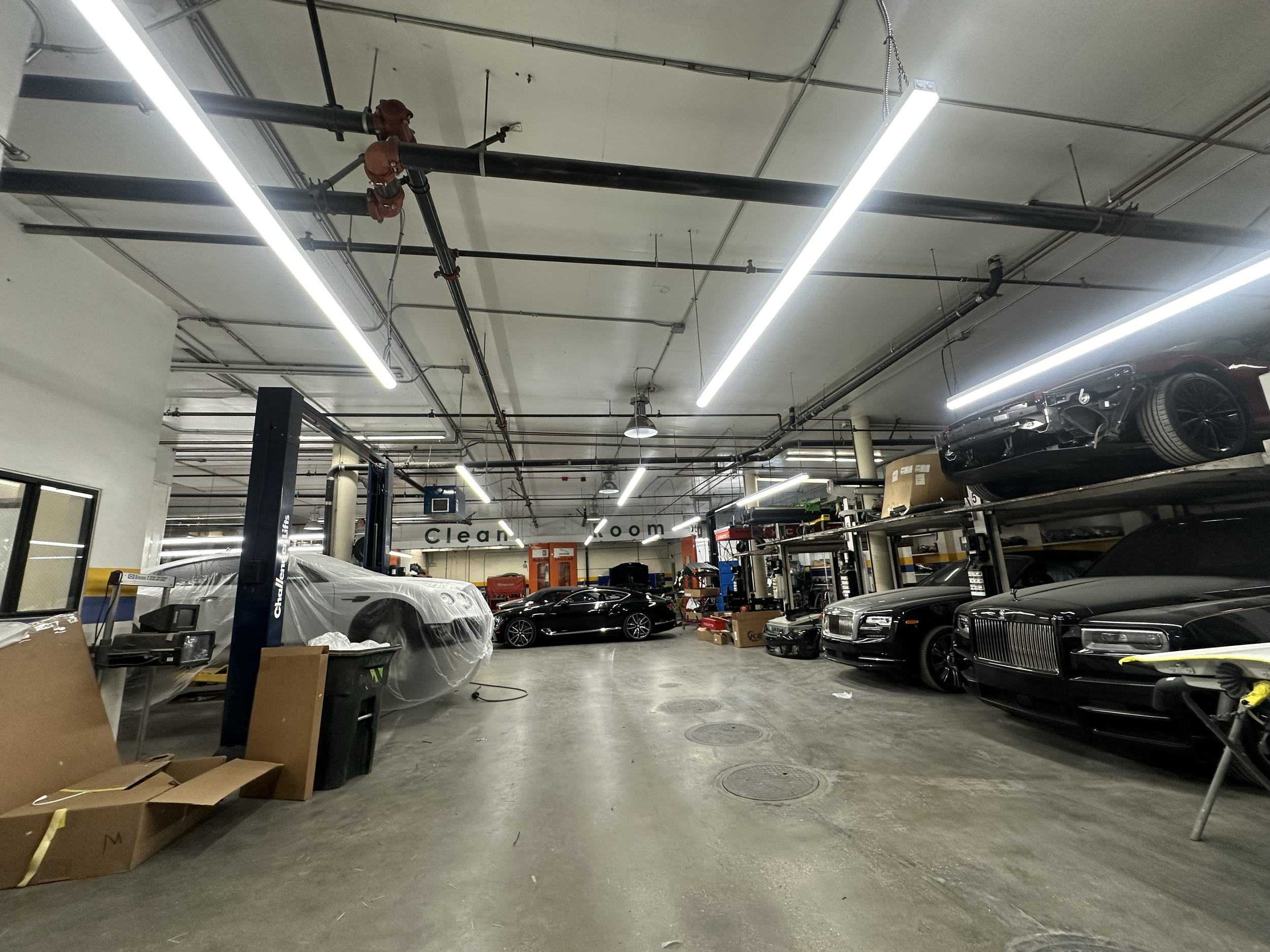 Indoor parking garage with several luxury cars parked, some covered with protective sheets. Overhead fluorescent lights, exposed piping on the ceiling, and boxes and miscellaneous items around.