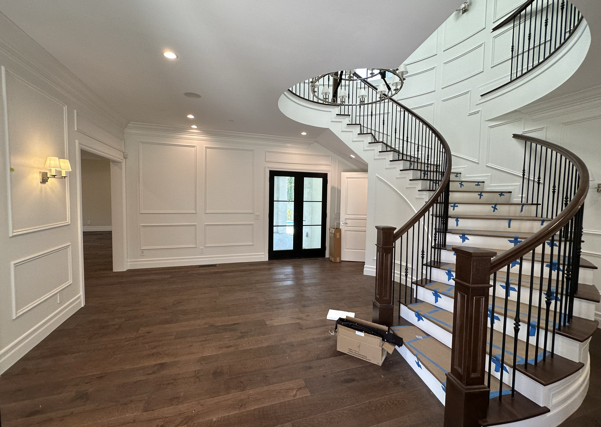 Interior of a house under renovation with a curved staircase, white paneled walls, hardwood floors, and a chandelier hanging from the ceiling. Blue painter's tape covers parts of the staircase steps, and there is a cardboard box and some tools on the