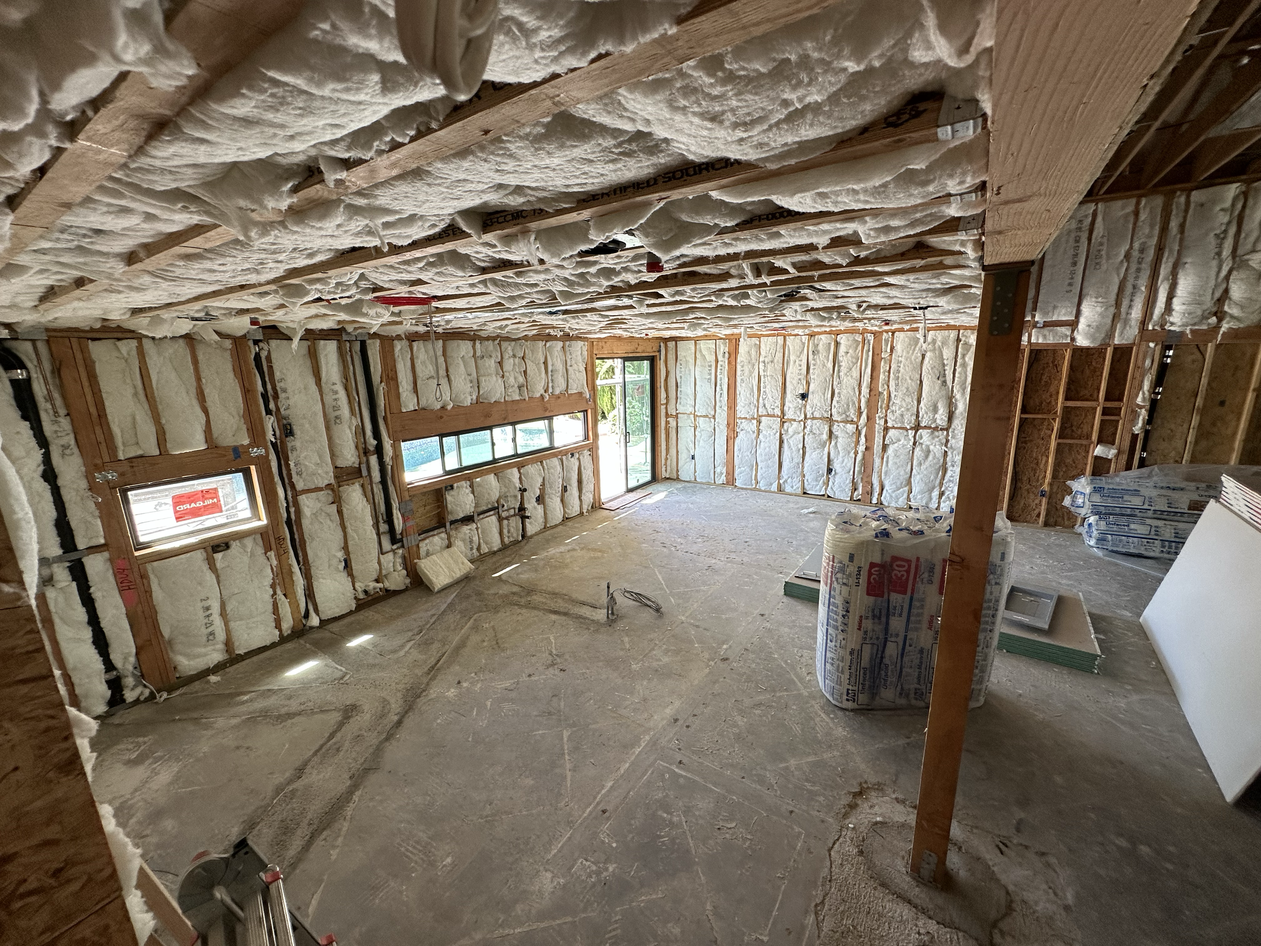 Interior view of a building under construction with insulation foam being installed between wooden wall studs. There are windows and construction supplies visible.