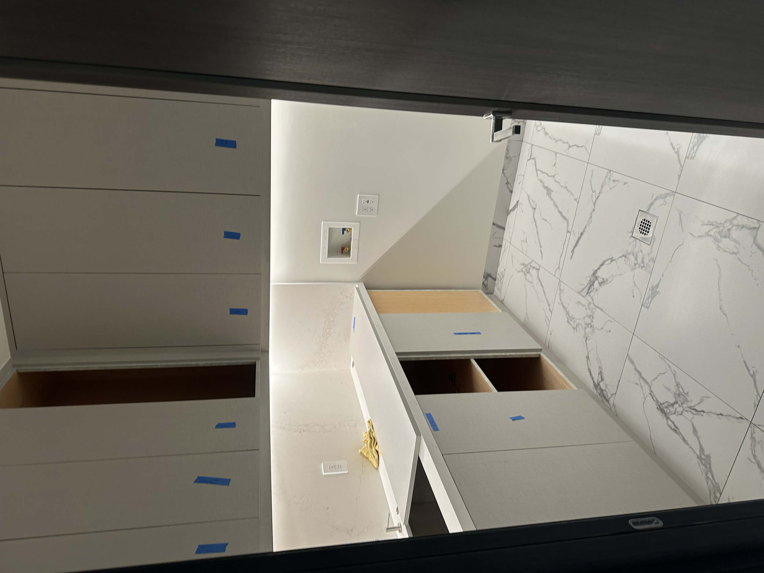An unfinished room with light-colored walls and marble tile flooring. Cabinets or closets line the left side, with some covered in blue painter's tape. There are electrical outlets and a vent on the wall, and a cardboard box and cloth on the floor ne