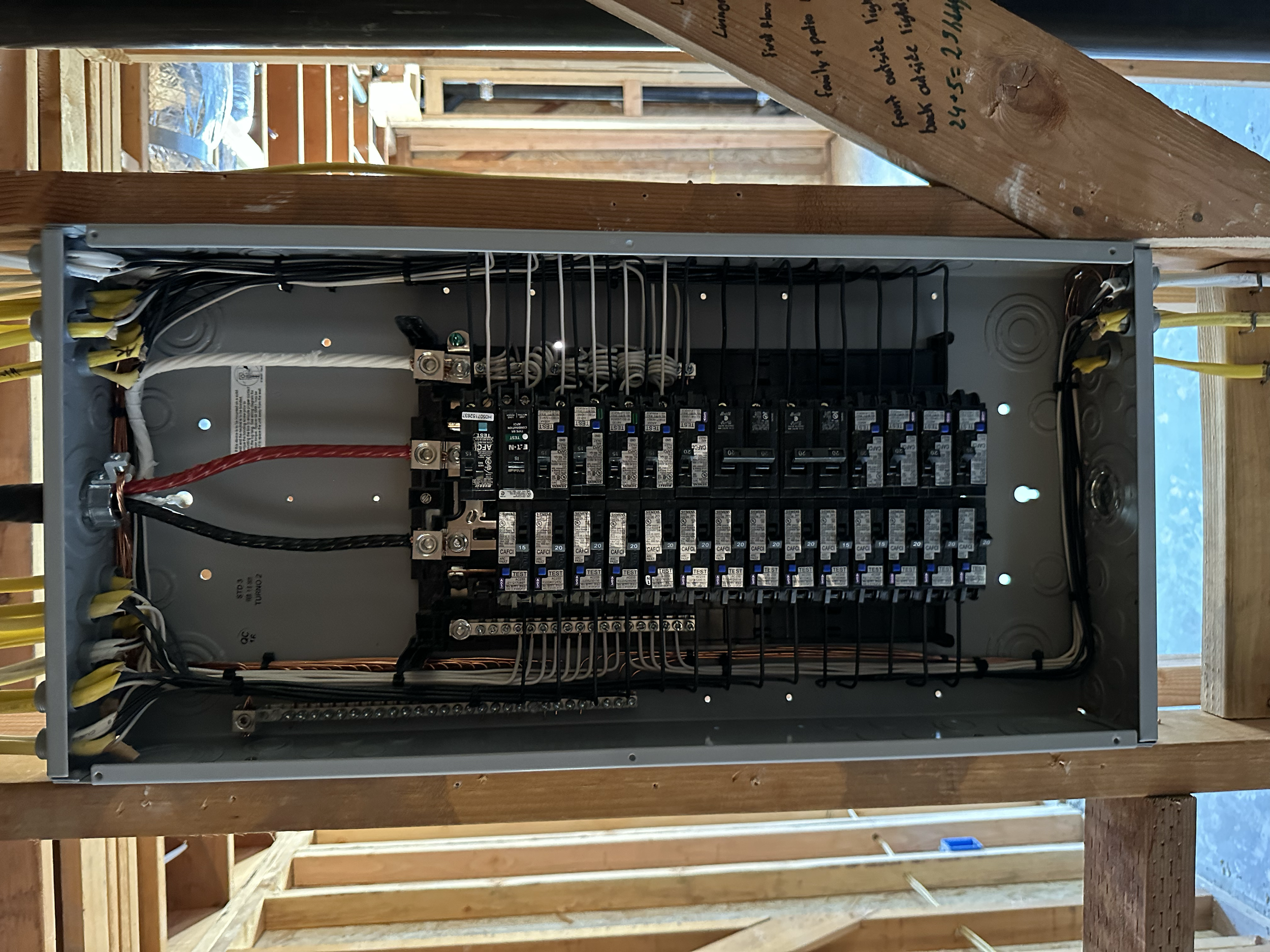 Electrical circuit breaker box with wires and labeled circuit breakers inside a wooden wall frame.