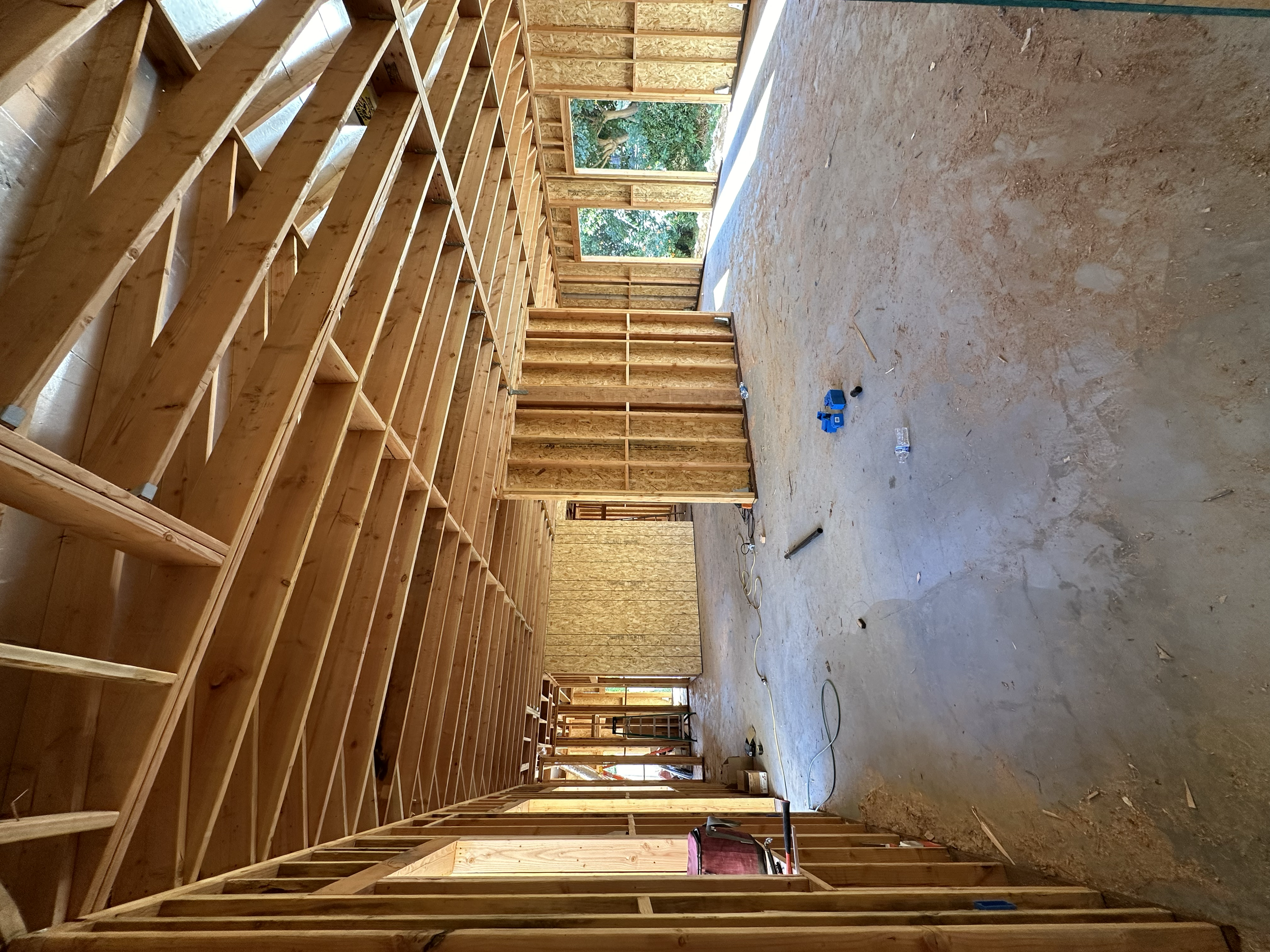 Interior view of a house under construction with exposed wooden framing and open wall studs, showing an unfinished room with a dirt floor, construction tools, and open doorways with a view of trees outside.