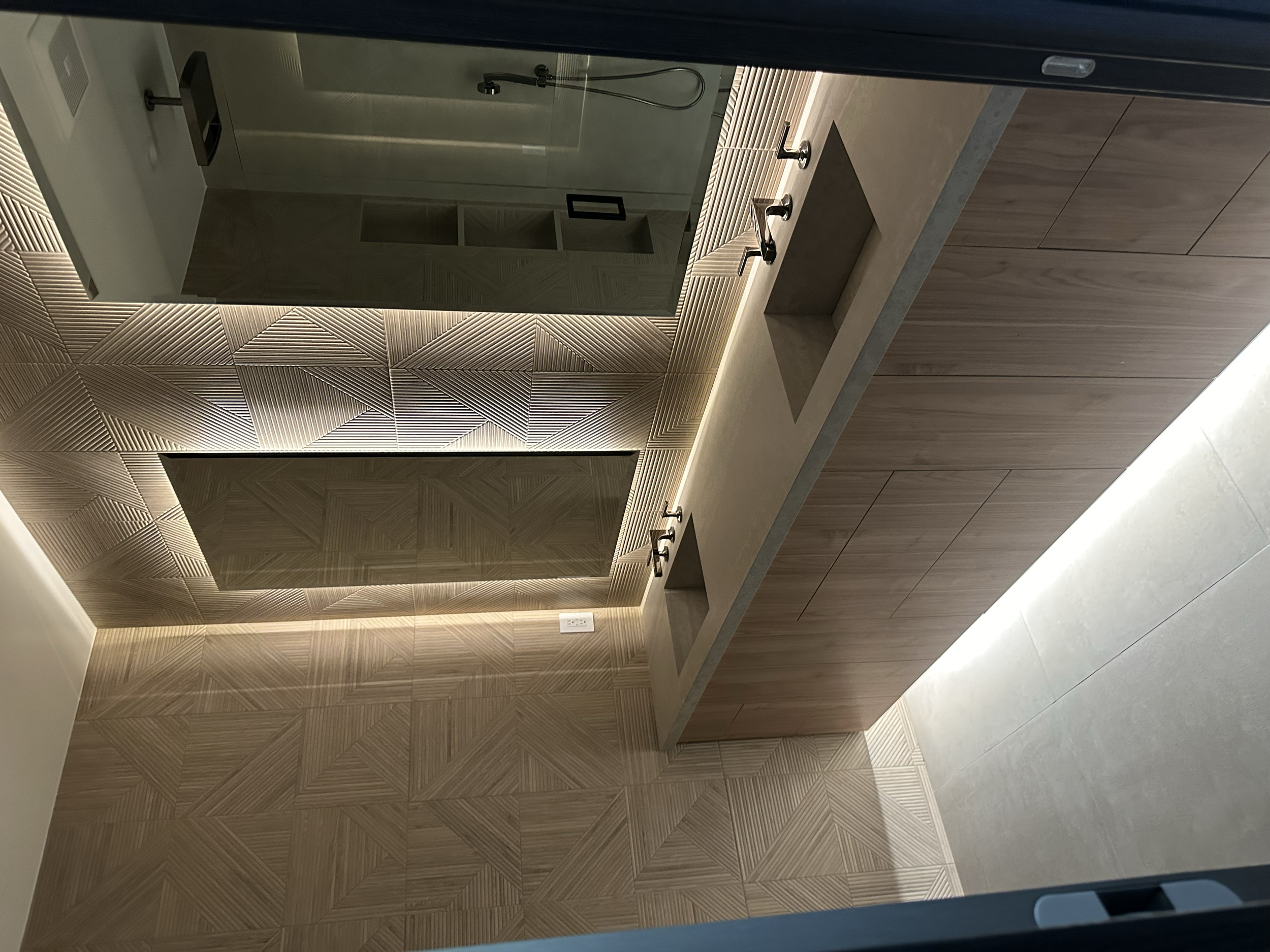 A modern bathroom with a shower enclosure featuring glass walls and a shower head, a long countertop with a built-in sink, and wooden cabinetry below. There is textured wall tiling with geometric patterns behind the shower.