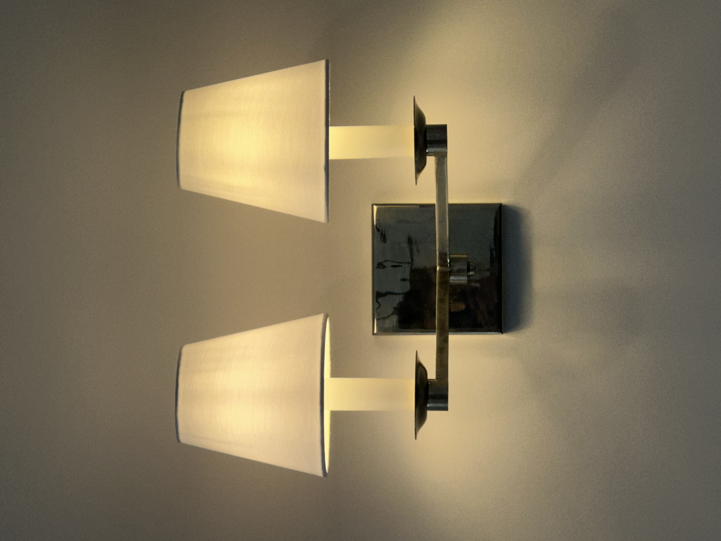 A modern wall-mounted lamp with a sleek design, featuring a rectangular fabric lampshade emitting warm light, attached to a metallic arm and wall plate.