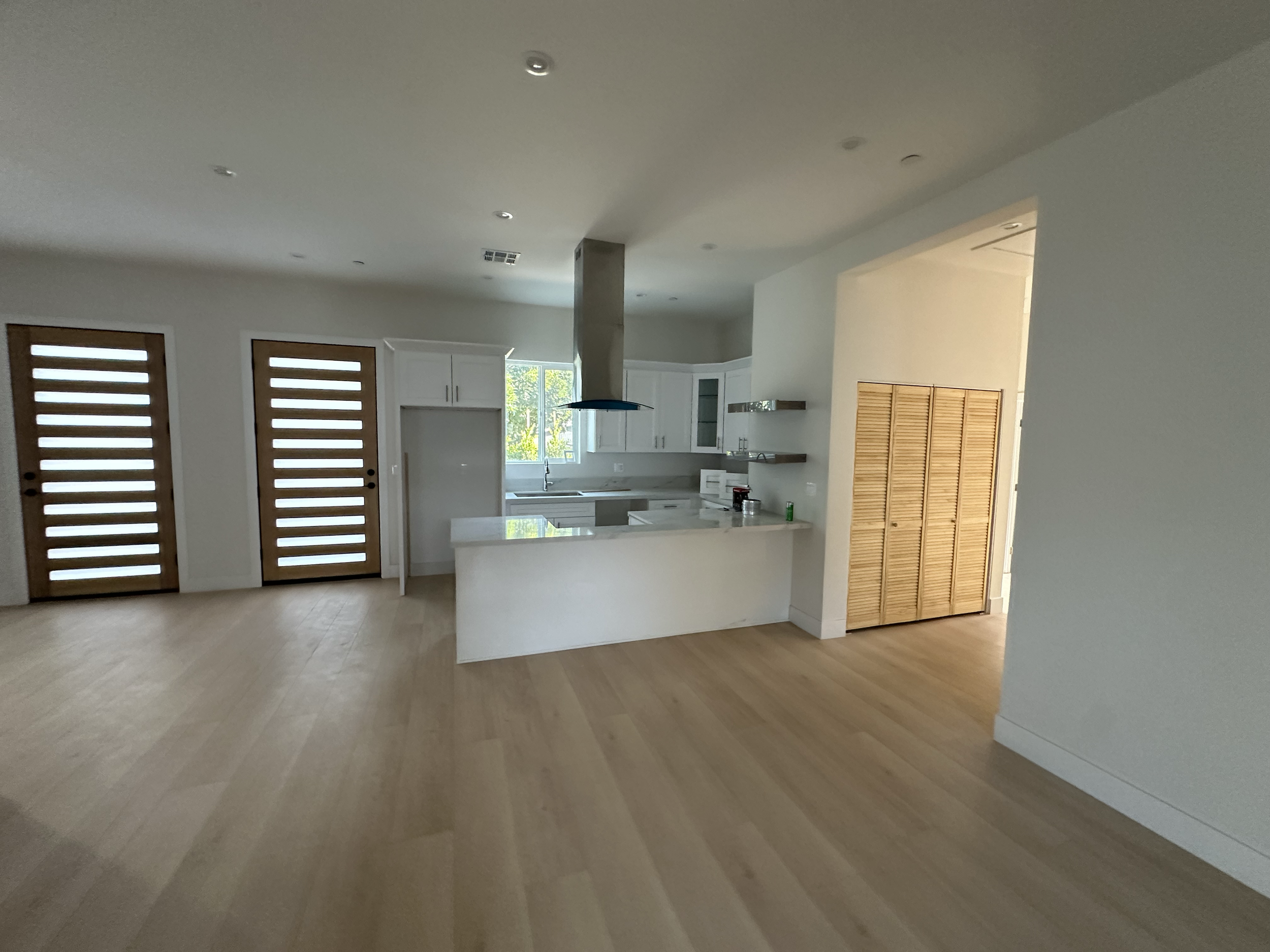 Interior view of a modern kitchen with white cabinets, a window, a kitchen hood, and wooden doors.