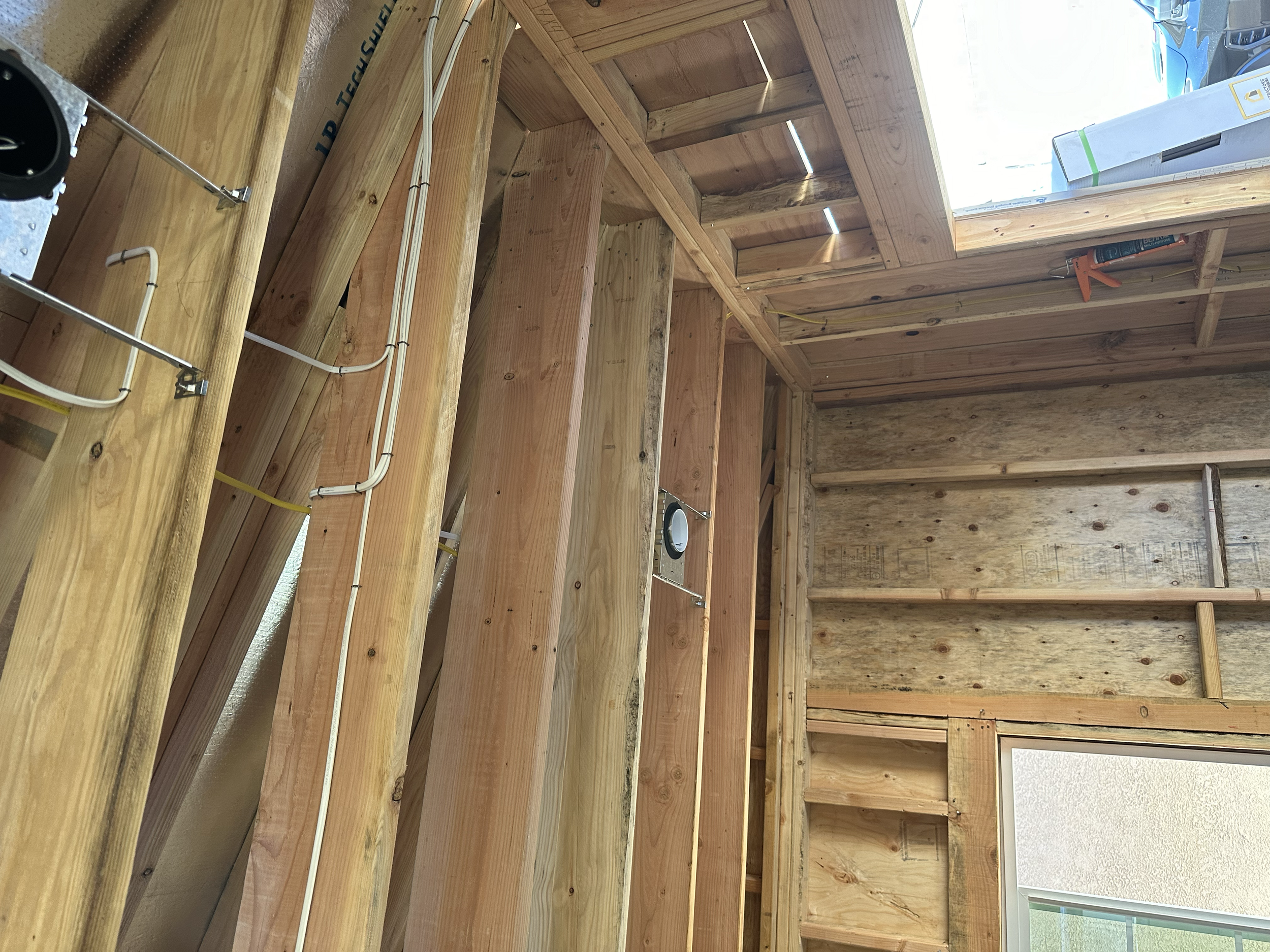 Inside a wooden structure, there are wooden framing and shelves, with some electrical wiring visible. Outside the window, a bright sky and some vehicles are seen.