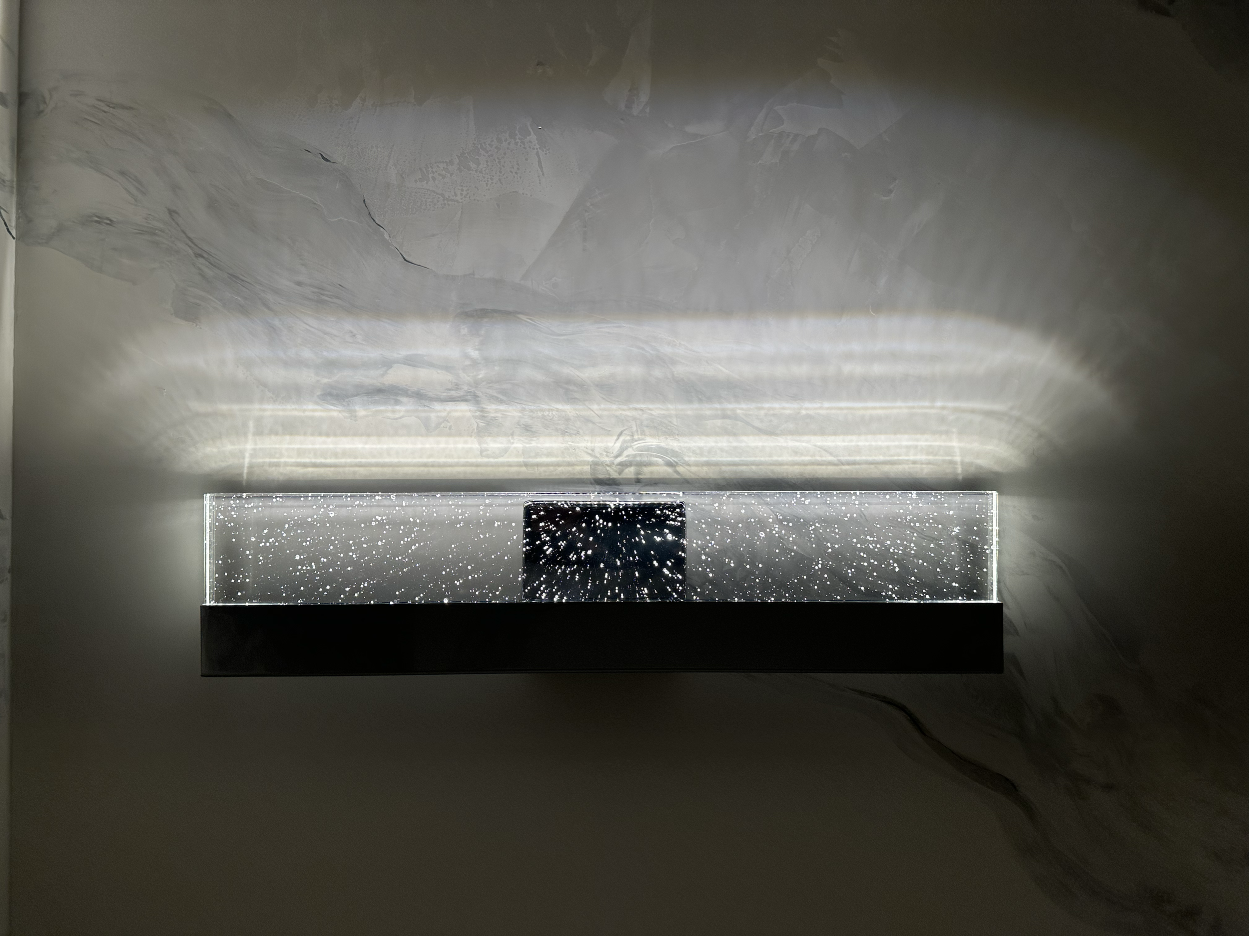 A modern, minimalist mirror with embedded LED lighting and speckled design, mounted on a marble wall illuminated by natural light from a nearby window.