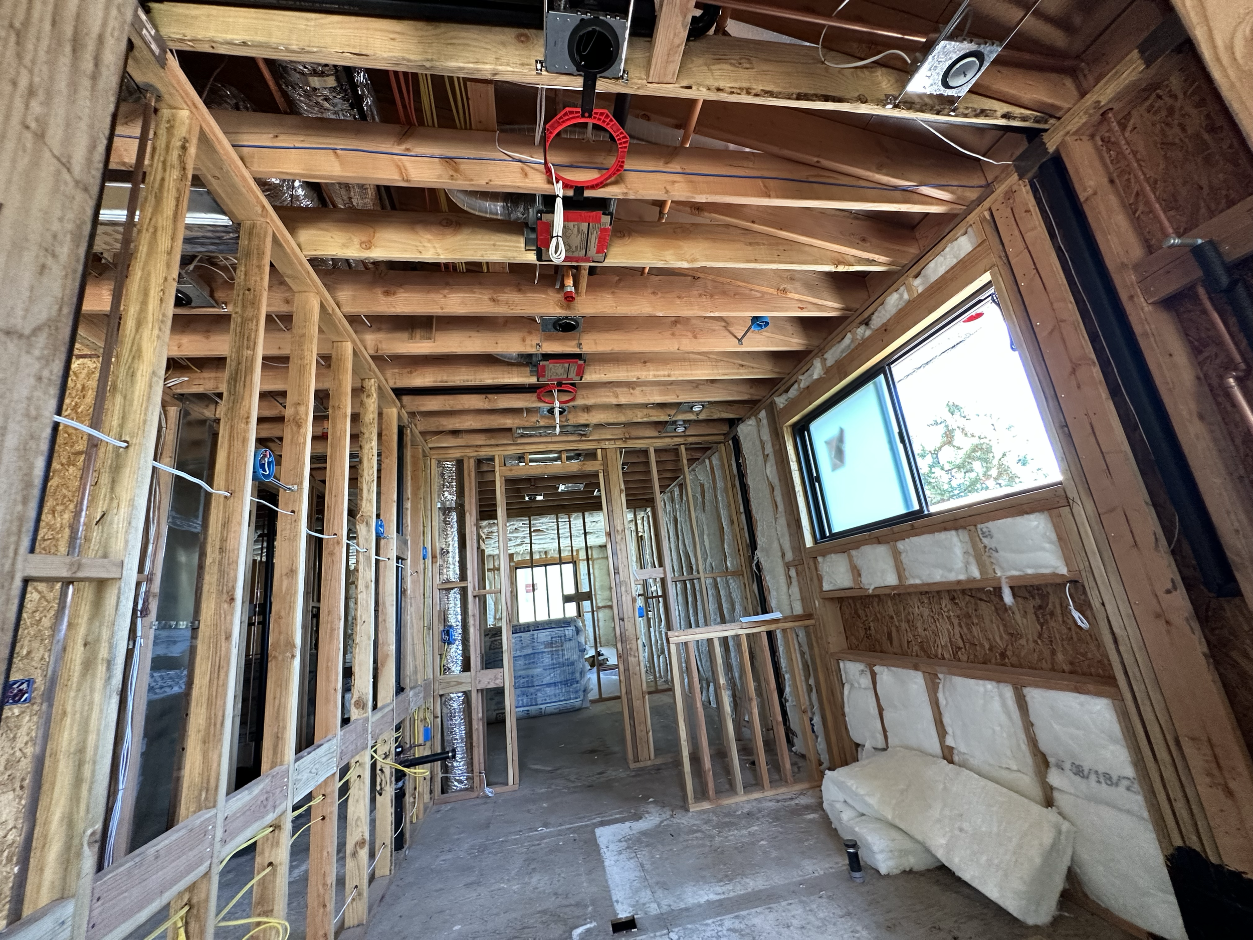 Interior view of a house under construction, showing exposed wooden framing, insulation, and electrical wiring, with a window on the right side.