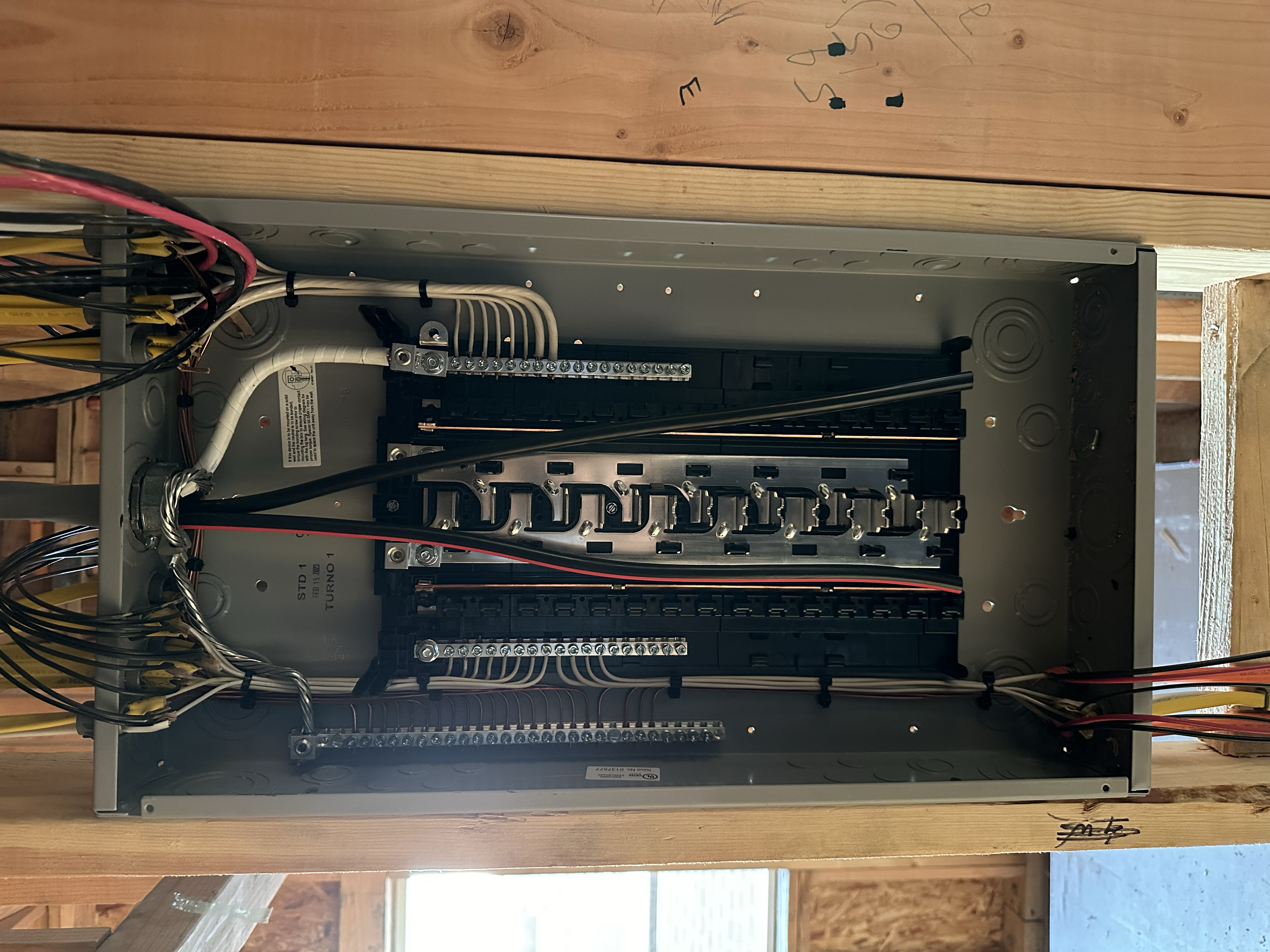 Open electrical panel with multiple wires, terminals, and circuit breakers, mounted on a wooden surface.