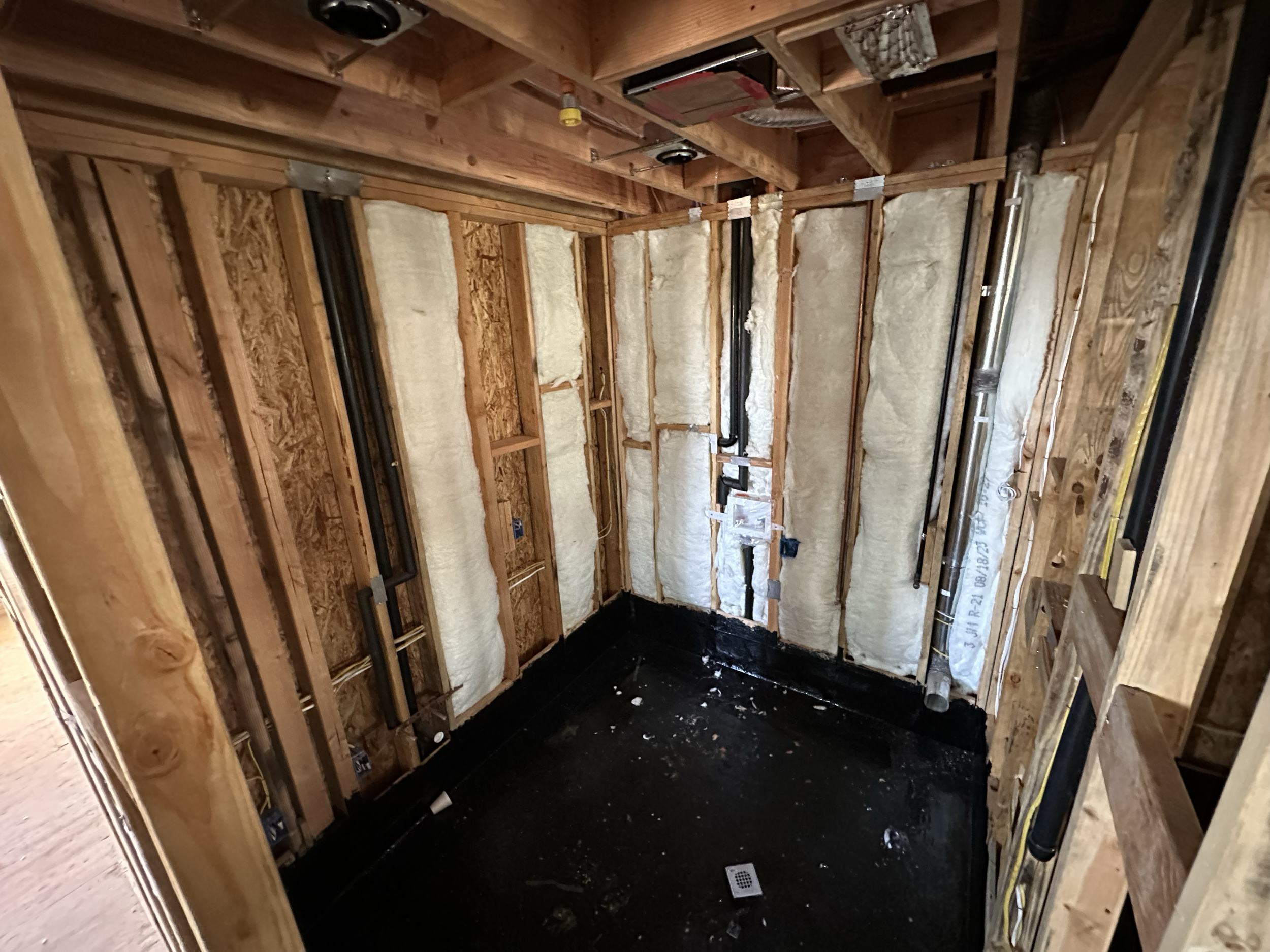 Inside view of a bathroom after demolition, showing wooden framing, insulation, plumbing pipes, and a black waterproofing layer on the floor.