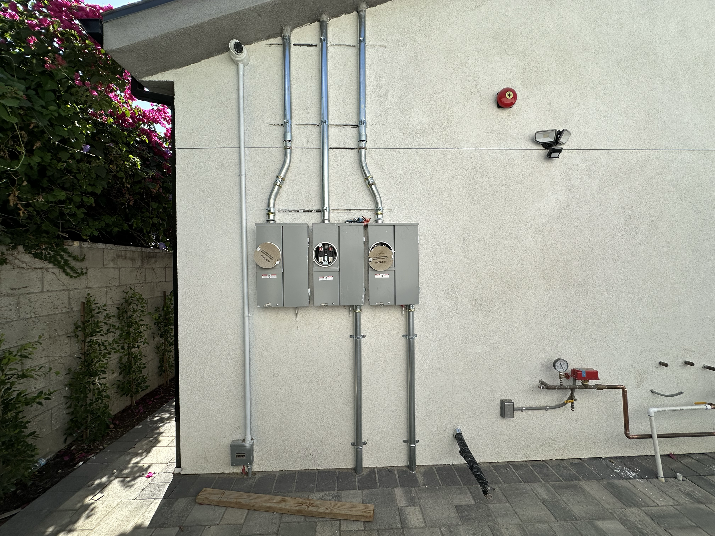 Exterior wall with electrical boxes, metal conduit pipes, security cameras, outdoor lighting, and a water pressure gauge, with pink flowering plant on the left.