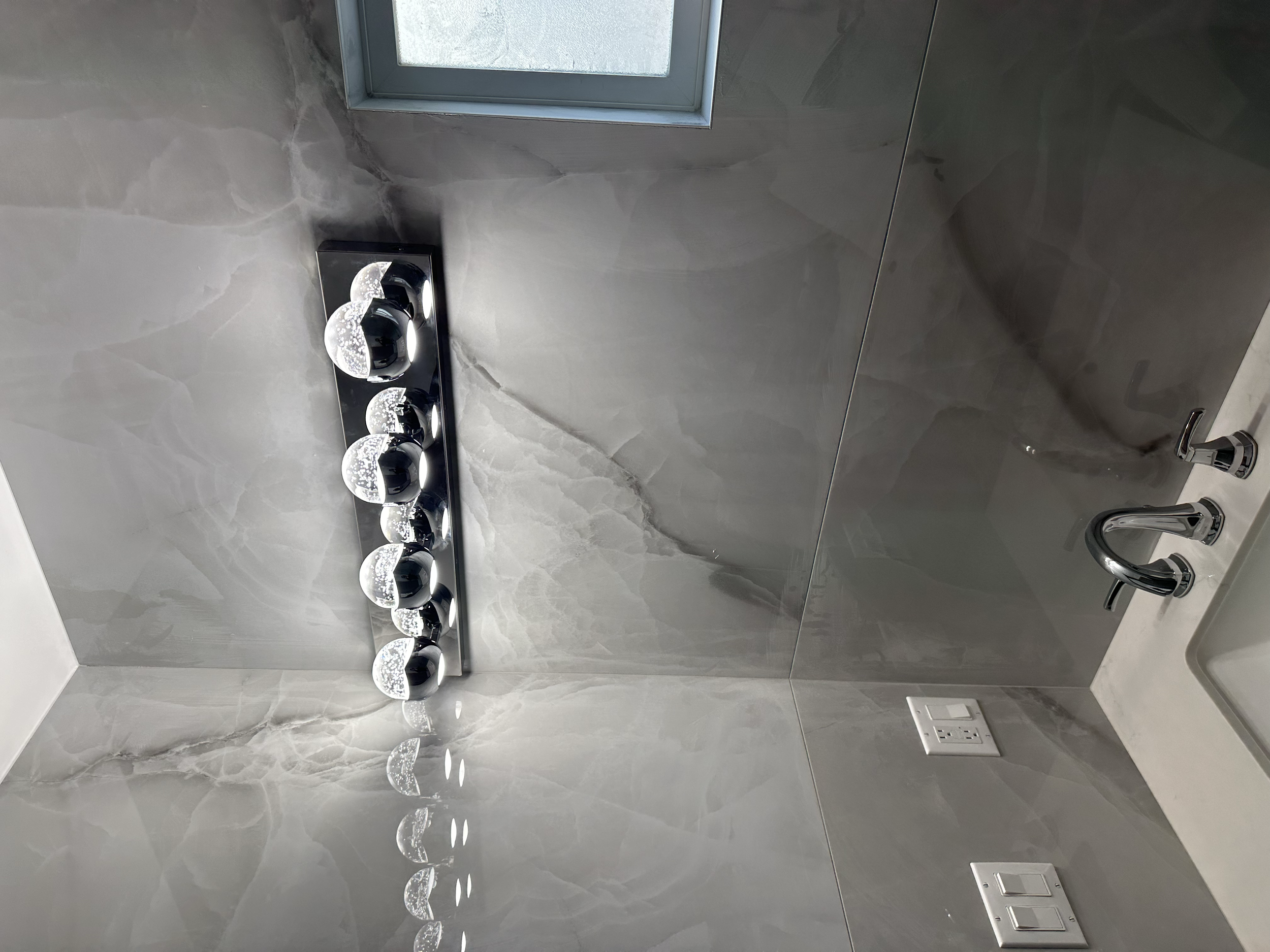 A section of a modern bathroom with grey marble walls, a window, a black horizontal light fixture with multiple bulbs, and a chrome faucet with a sink visible at the bottom right, and electrical outlets on the wall.
