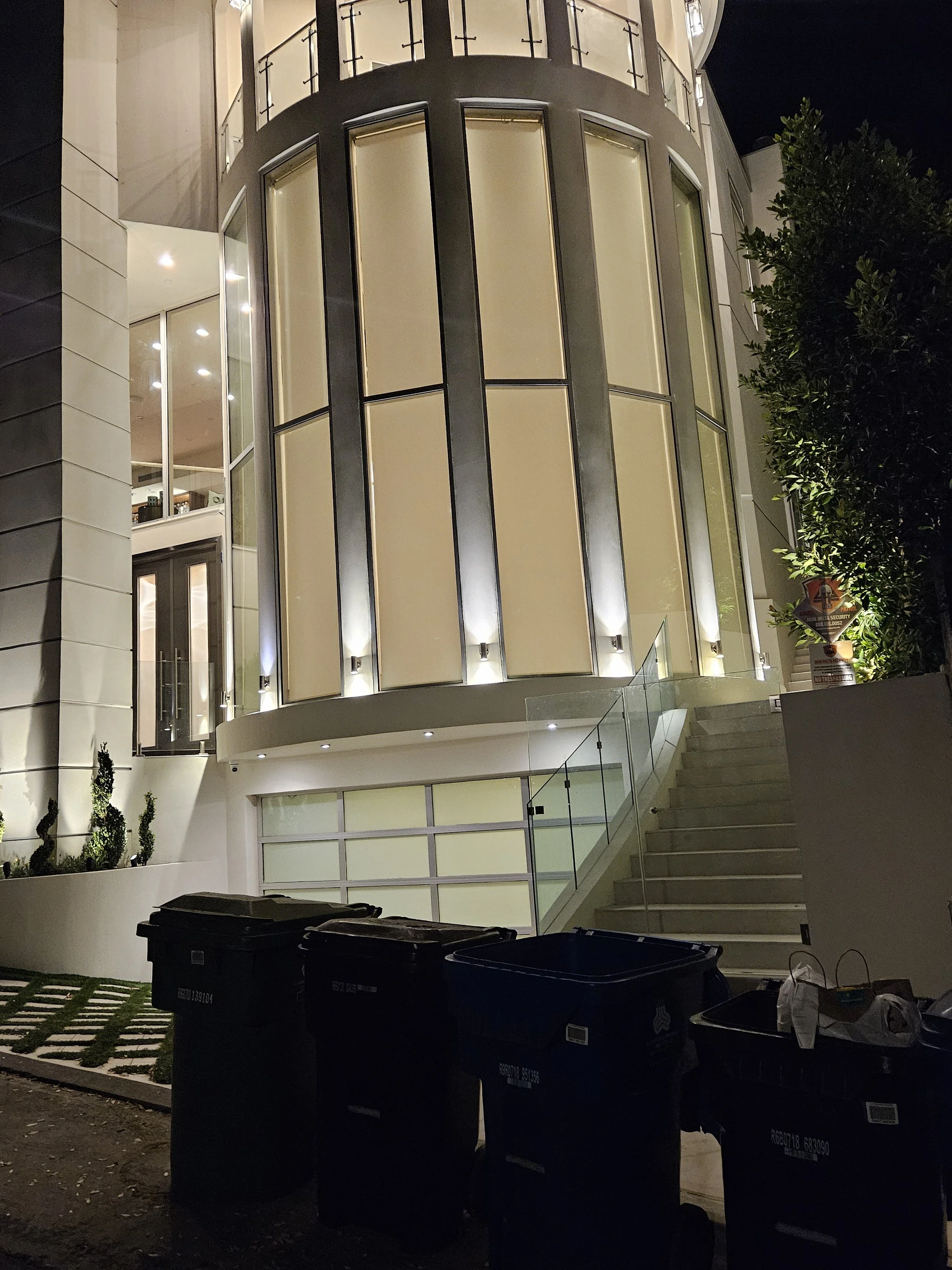 Nighttime view of a modern house with large illuminated windows and a staircase outside. Multiple trash bins are placed in front of the house.