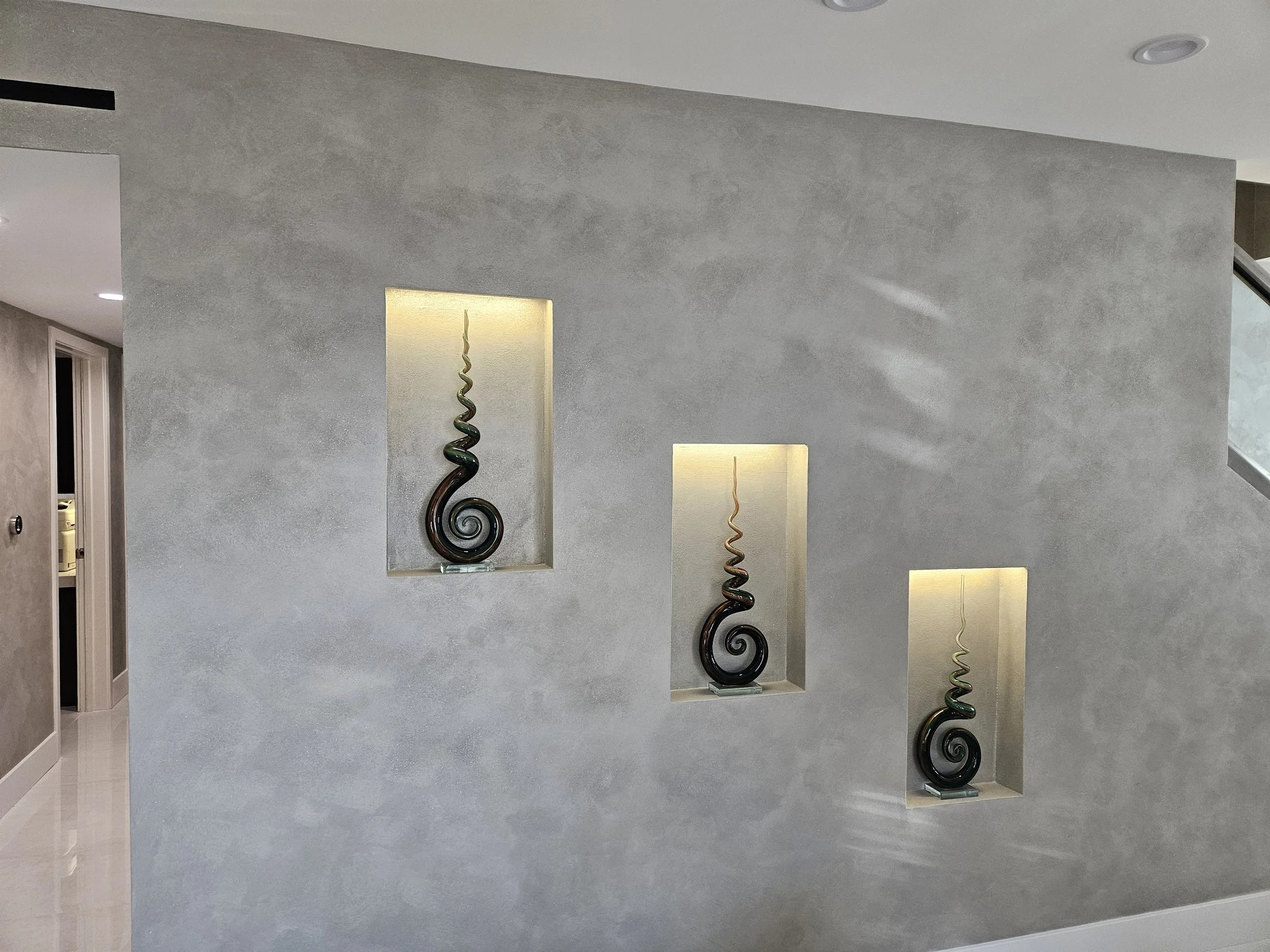 Interior of a modern house featuring recessed wall niches illuminated with warm light and containing decorative spiral sculptures, with a polished floor and a door on the left.