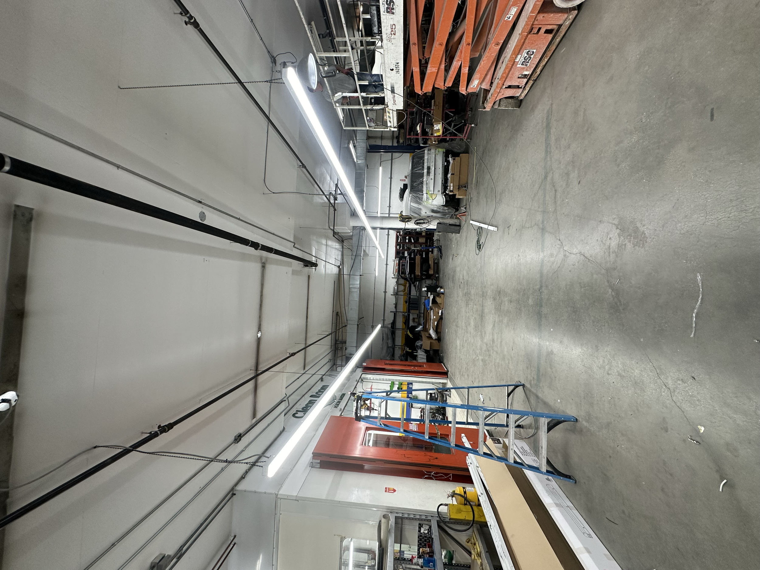 Industrial workspace with various equipment, tools, and scaffolding, including a bright lighting fixture, shelves with supplies, and workspace tables.