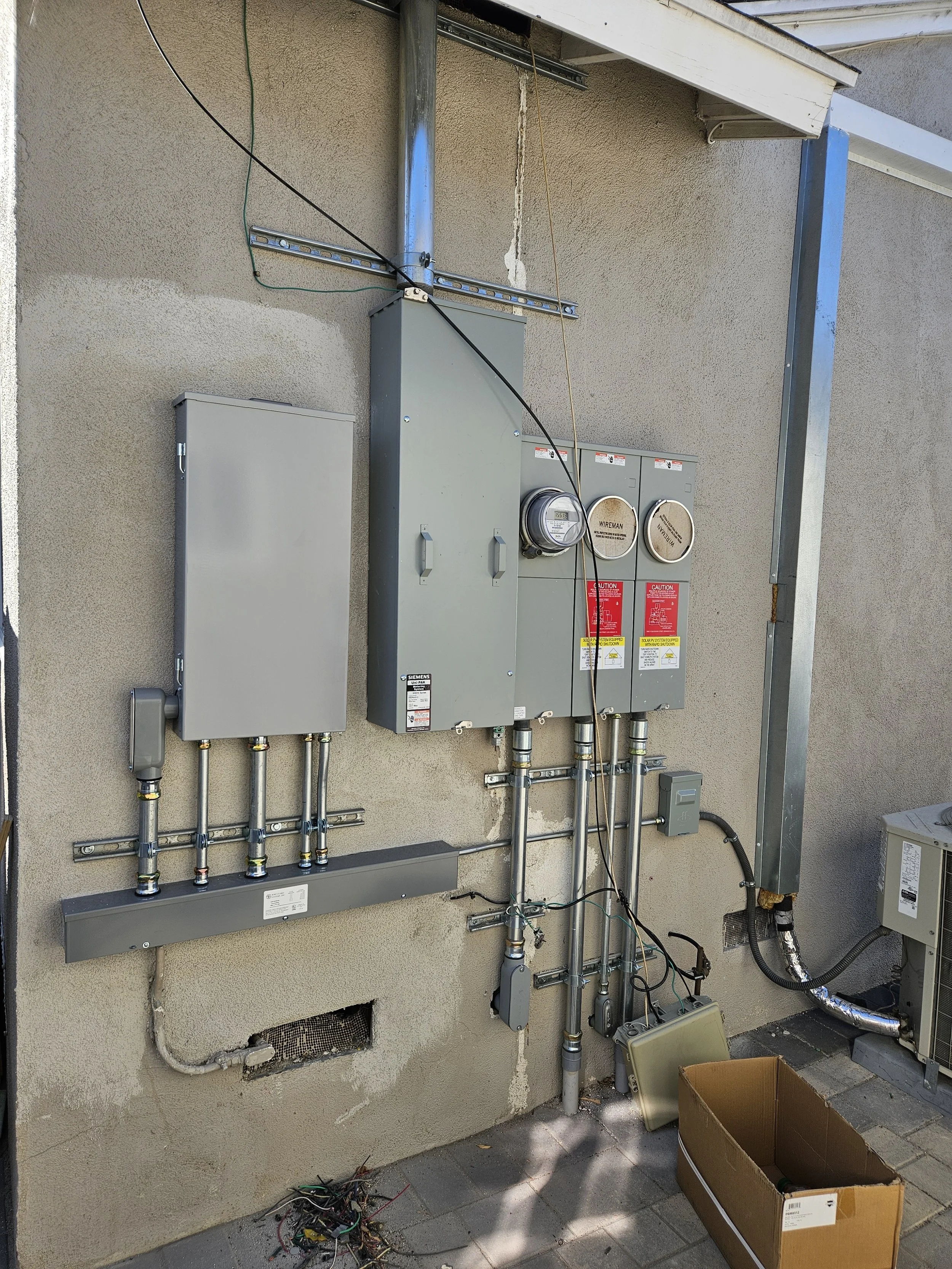 Outdoor utility area with electrical panels, wiring, and a vent pipe, some wiring on the ground, and an open cardboard box.