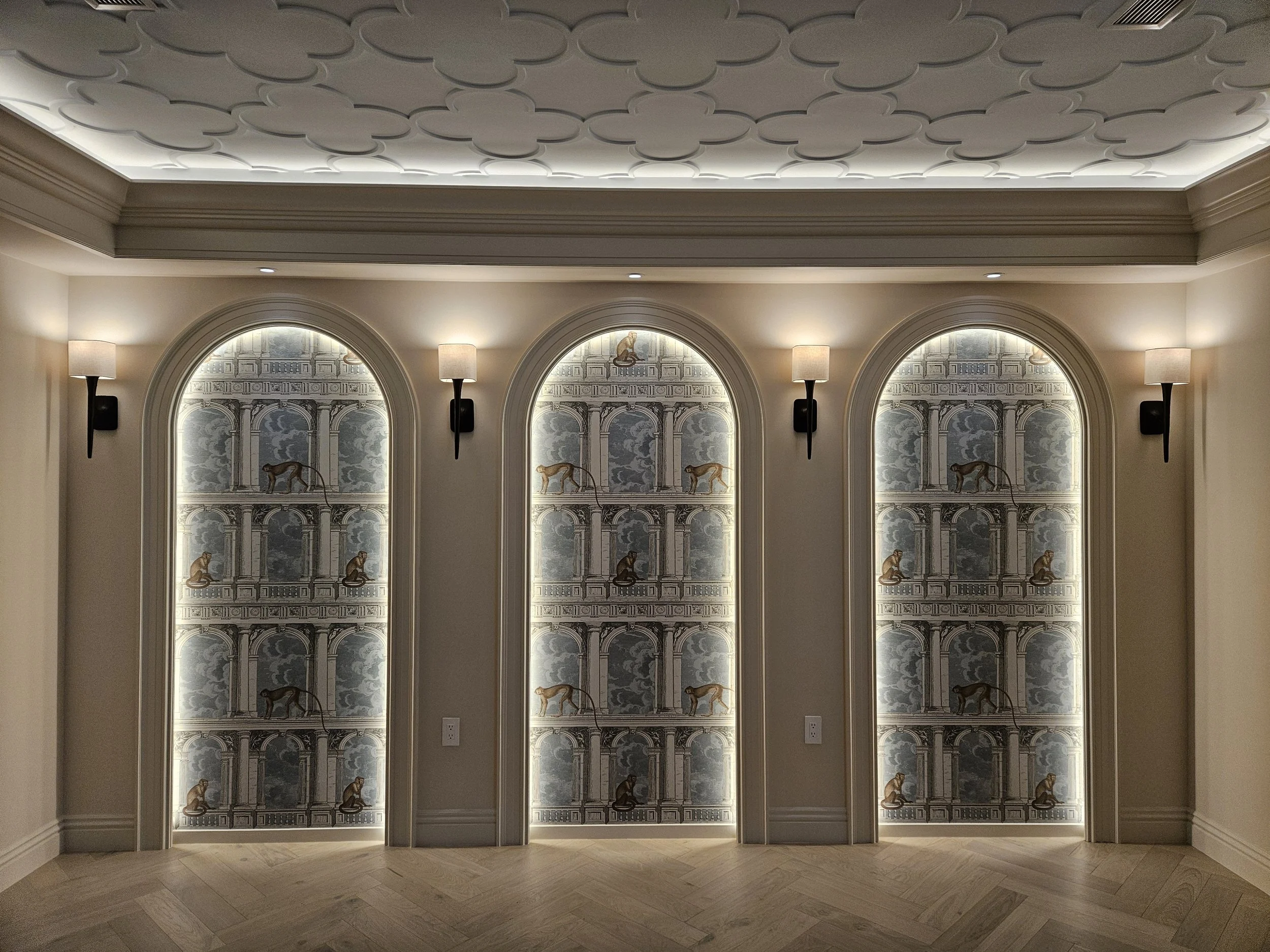 An interior wall with three large arched murals depicting architectural scenes with animals, illuminated by wall sconces, with a coffered ceiling above.