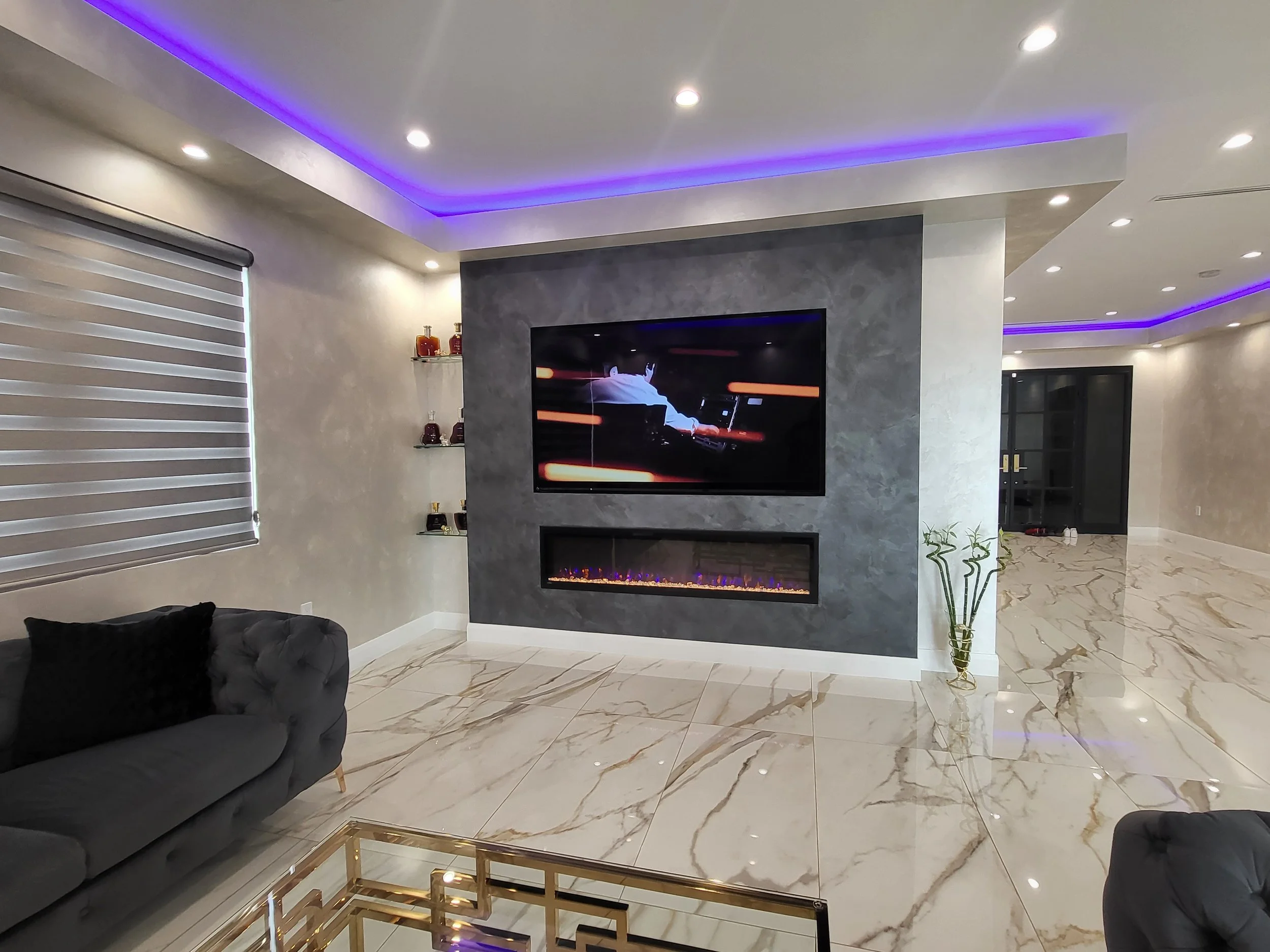 Modern living room with marble flooring, a sleek black sofa, a wall-mounted TV, a fireplace, and purple LED lighting accents on the ceiling.