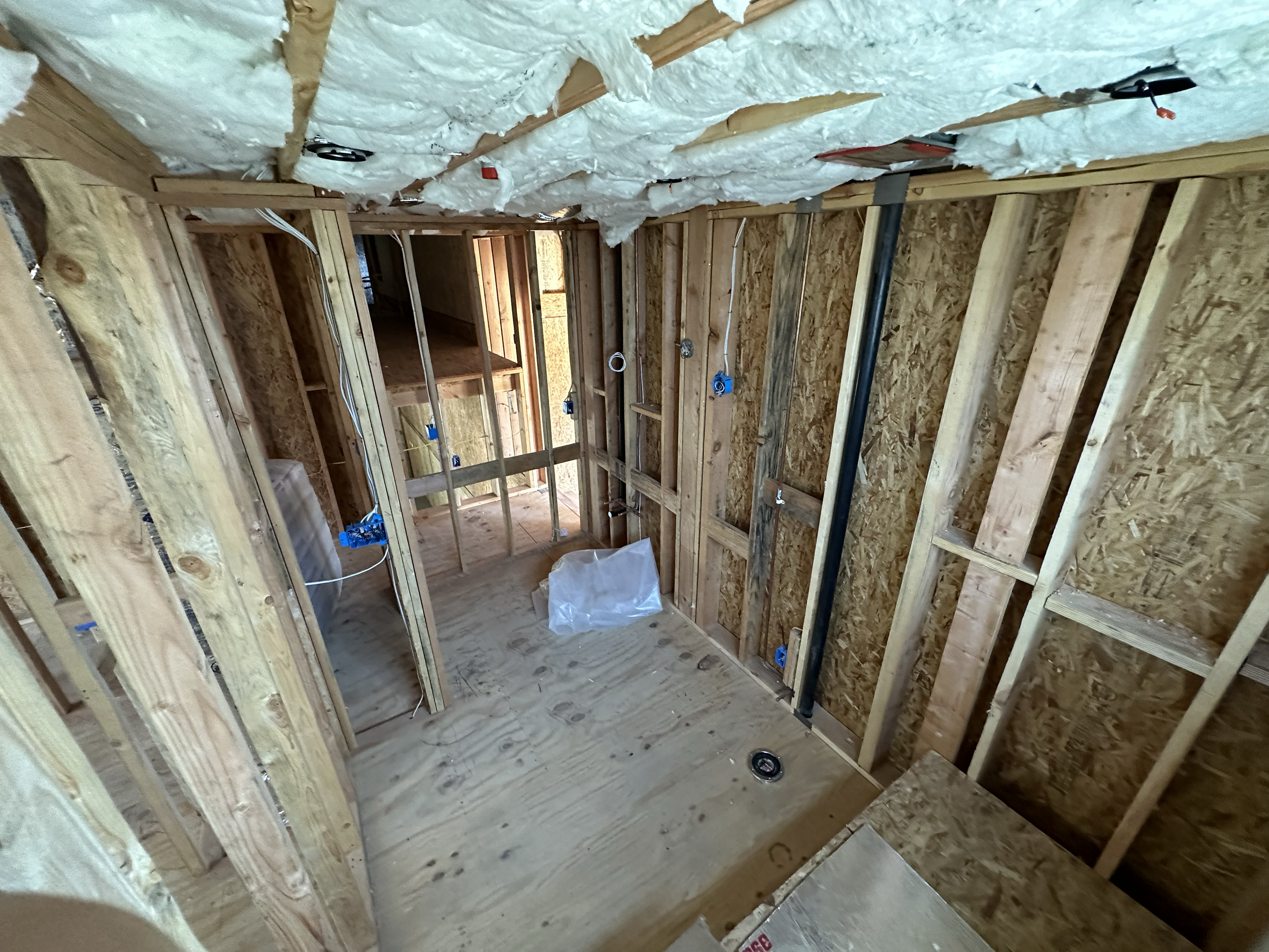 Interior view of a building under construction with exposed wooden framework, insulation in ceiling, electrical wiring, and plumbing pipes visible.