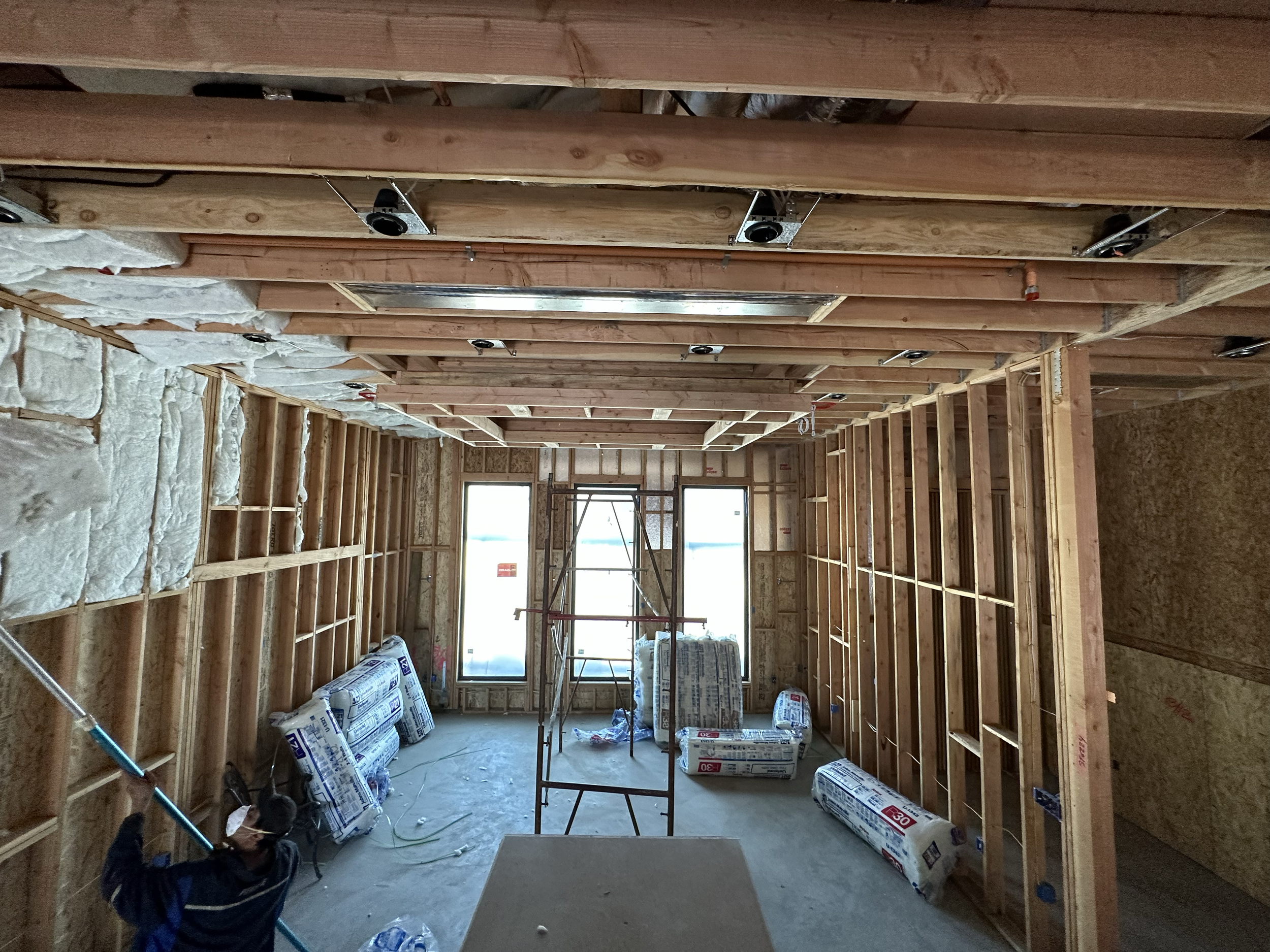 Interior of a building under construction with exposed wooden framing, insulation, and construction materials on the floor, with windows at the far end.
