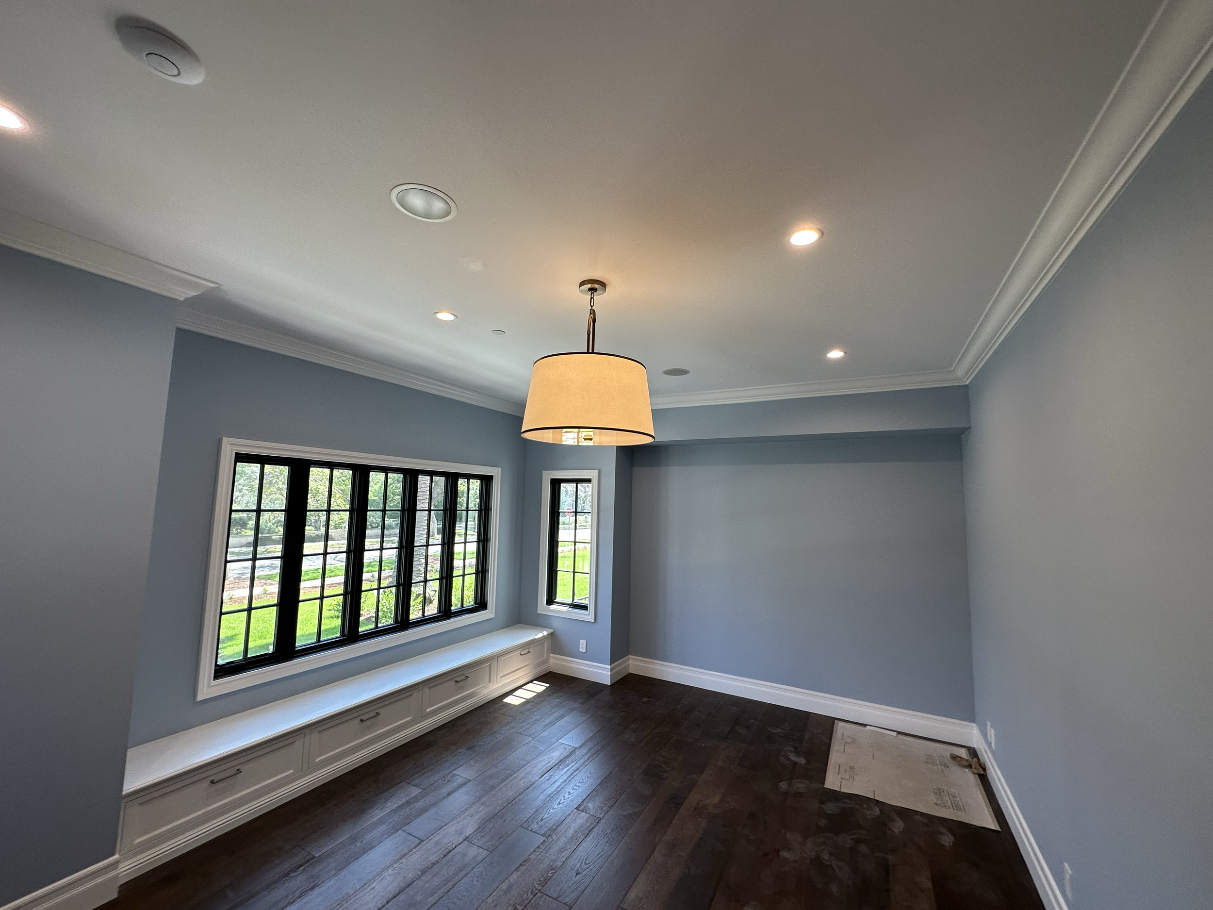 Living room with light blue walls, large black-framed windows, a built-in white window seat, hardwood flooring, and a ceiling with installed lights and a hanging lamp.