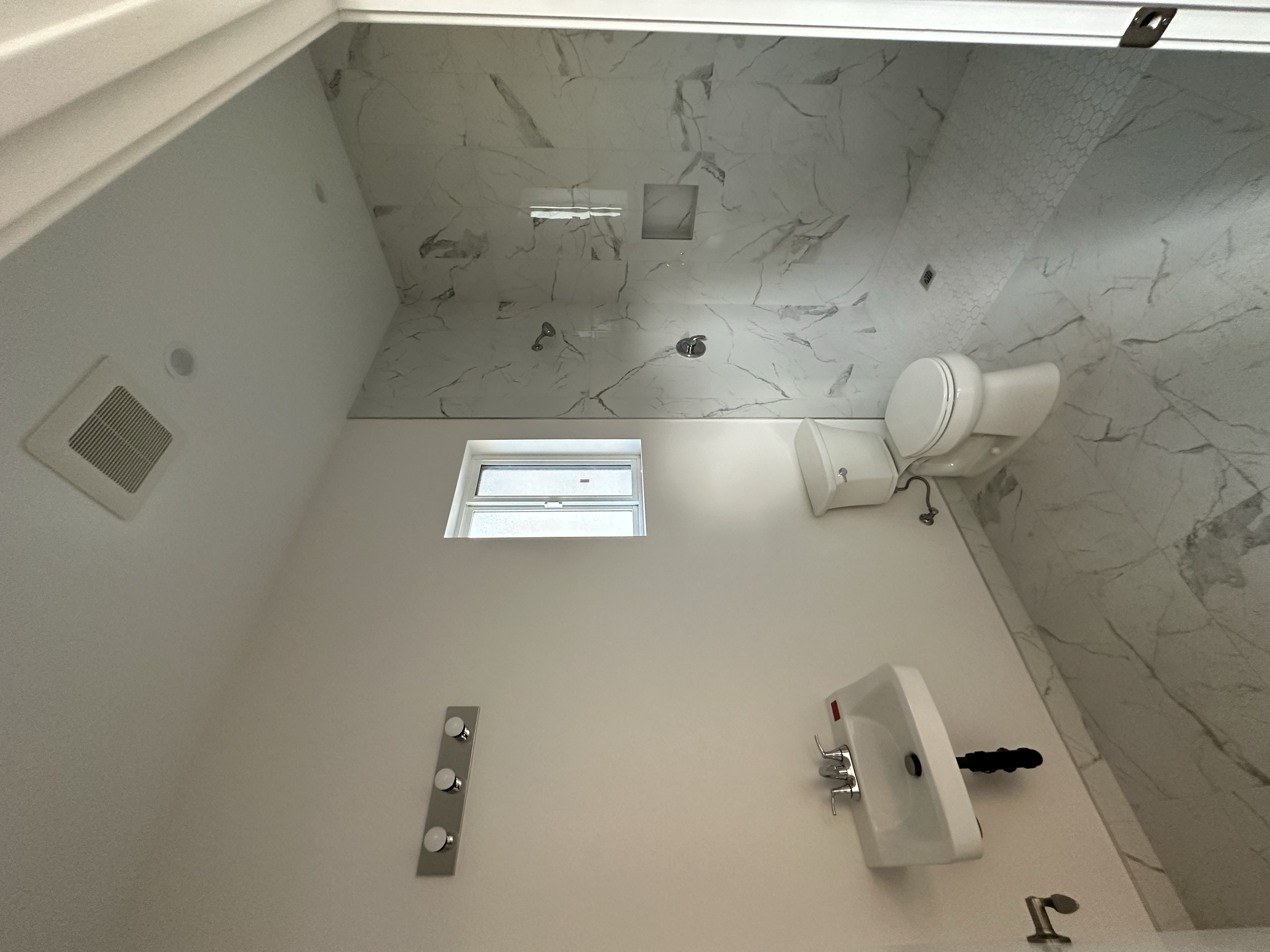 A bathroom with white marble walls and floor, a window, a toilet, a mirror with lights, and a sink.