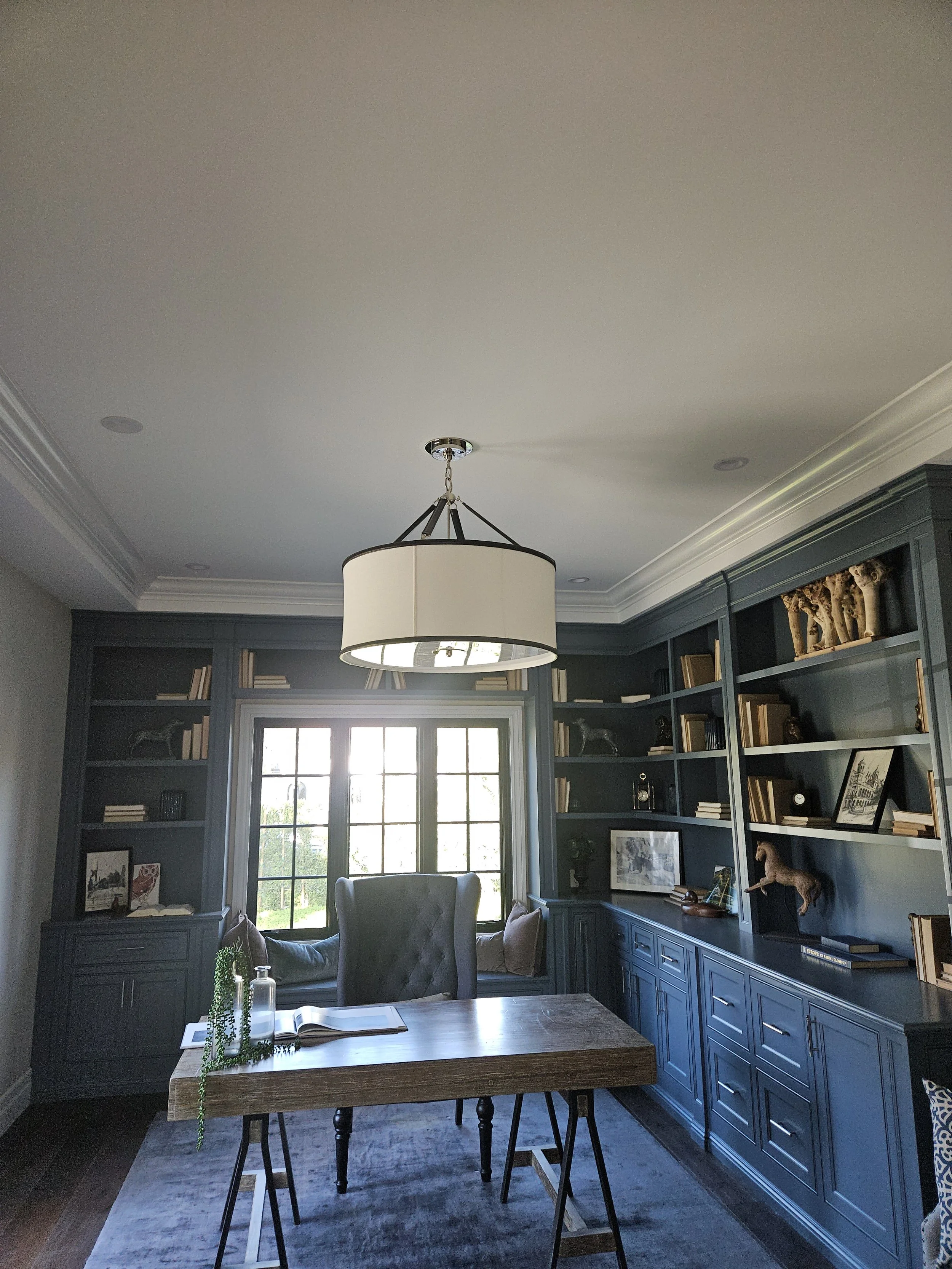 A home office with built-in blue bookshelves filled with books and decorative items, large window allowing natural light, a wooden table with a book and a glass bottle with a green plant, a blue rug, a gray upholstered chair, and a circular ceiling l