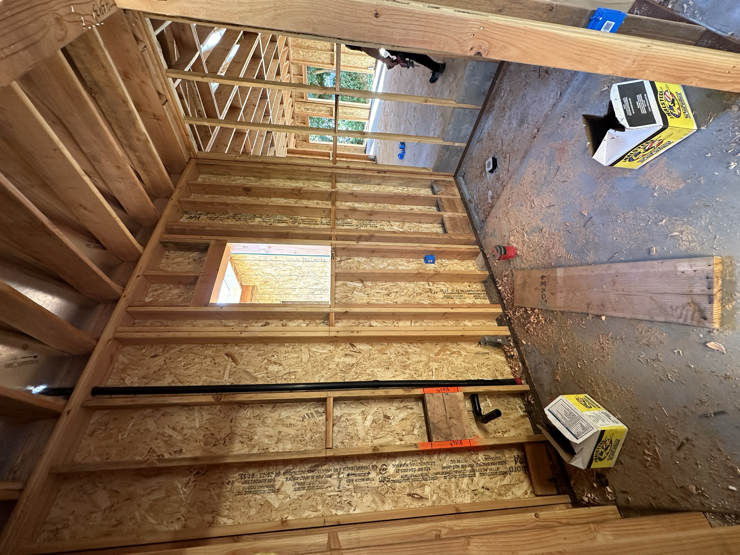 The interior of a building under construction with wooden framing, exposed OSB walls, construction tools, and materials on the floor.