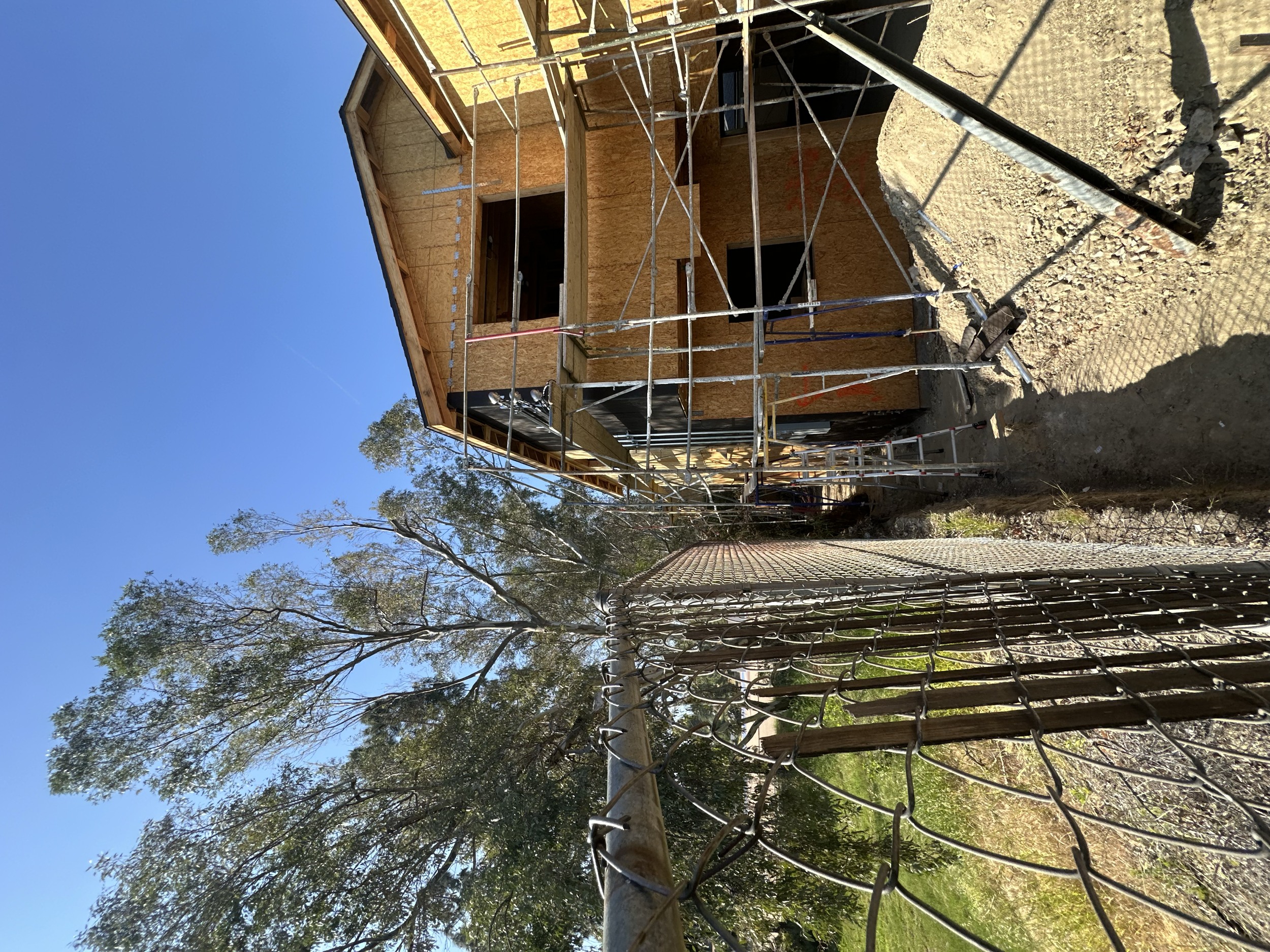 A house under construction with scaffolding around it, located next to a chain-link fence, with trees in the background and a clear blue sky.