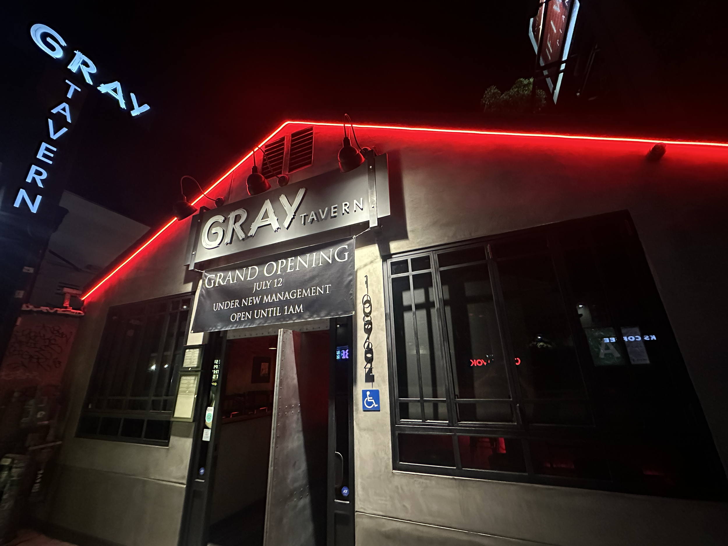 Night exterior of Gray Tavern with neon sign, red neon outlining the building, and a sign announcing grand opening on July 12, with work under new management until 1AM, and accessibility disabled parking sign.