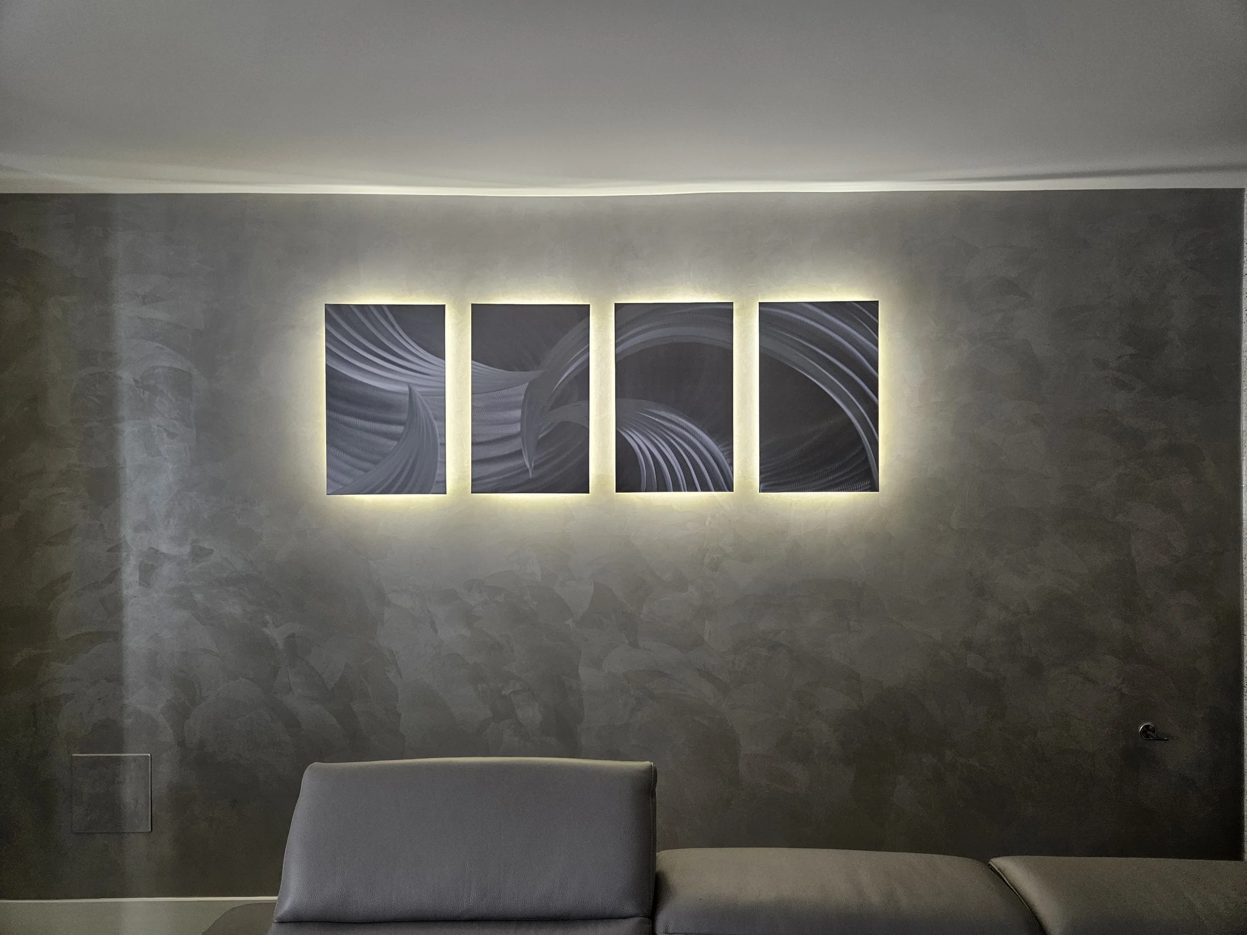 A modern interior wall with three abstract art pieces illuminated by backlighting, with a chair visible at the bottom of the image.