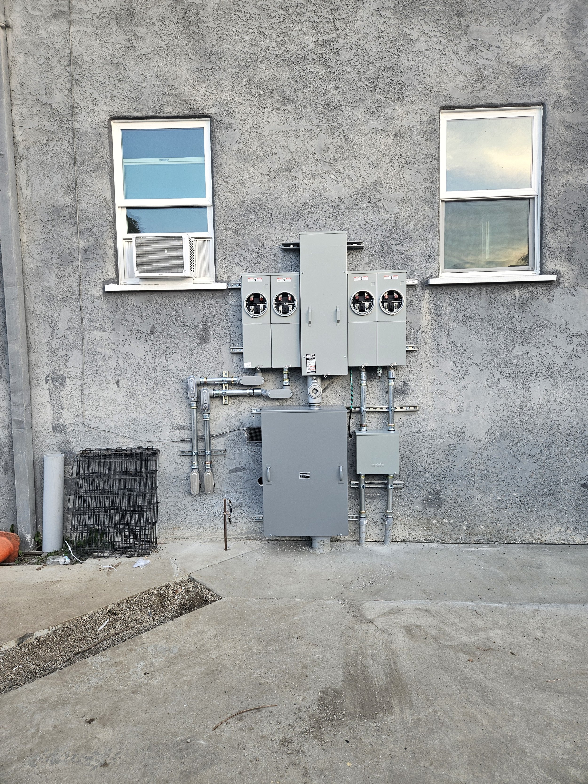 The image shows a gray textured exterior wall of a building with four windows and electrical/utility boxes mounted on the wall. The windows have frames, and two have air conditioning units. Below the wall is a concrete ground with some debris and con