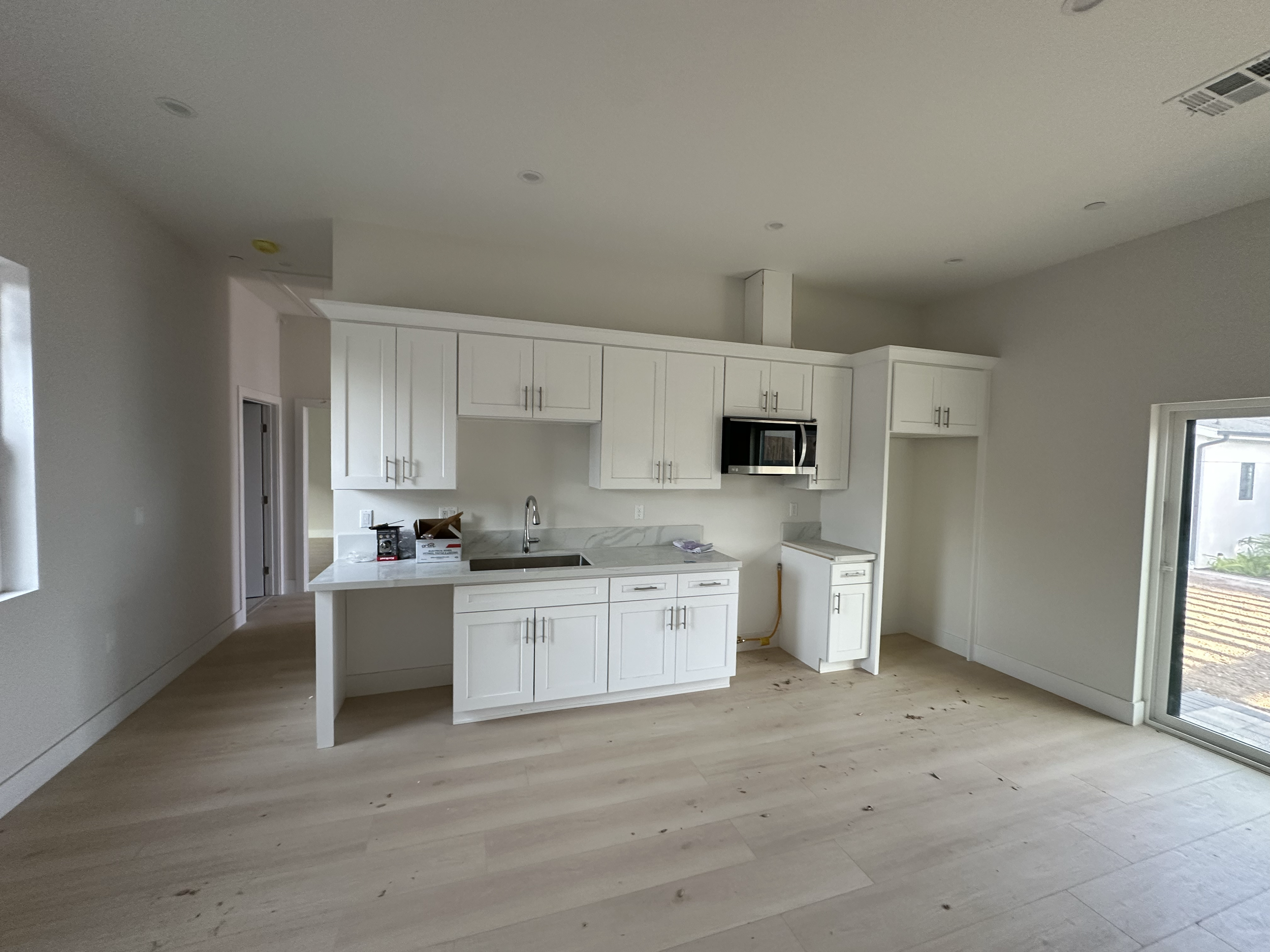 Unfinished kitchen with white cabinets, a marble countertop, stainless steel microwave, and light wood flooring. An open door leads outside, and there is construction debris on the floor.