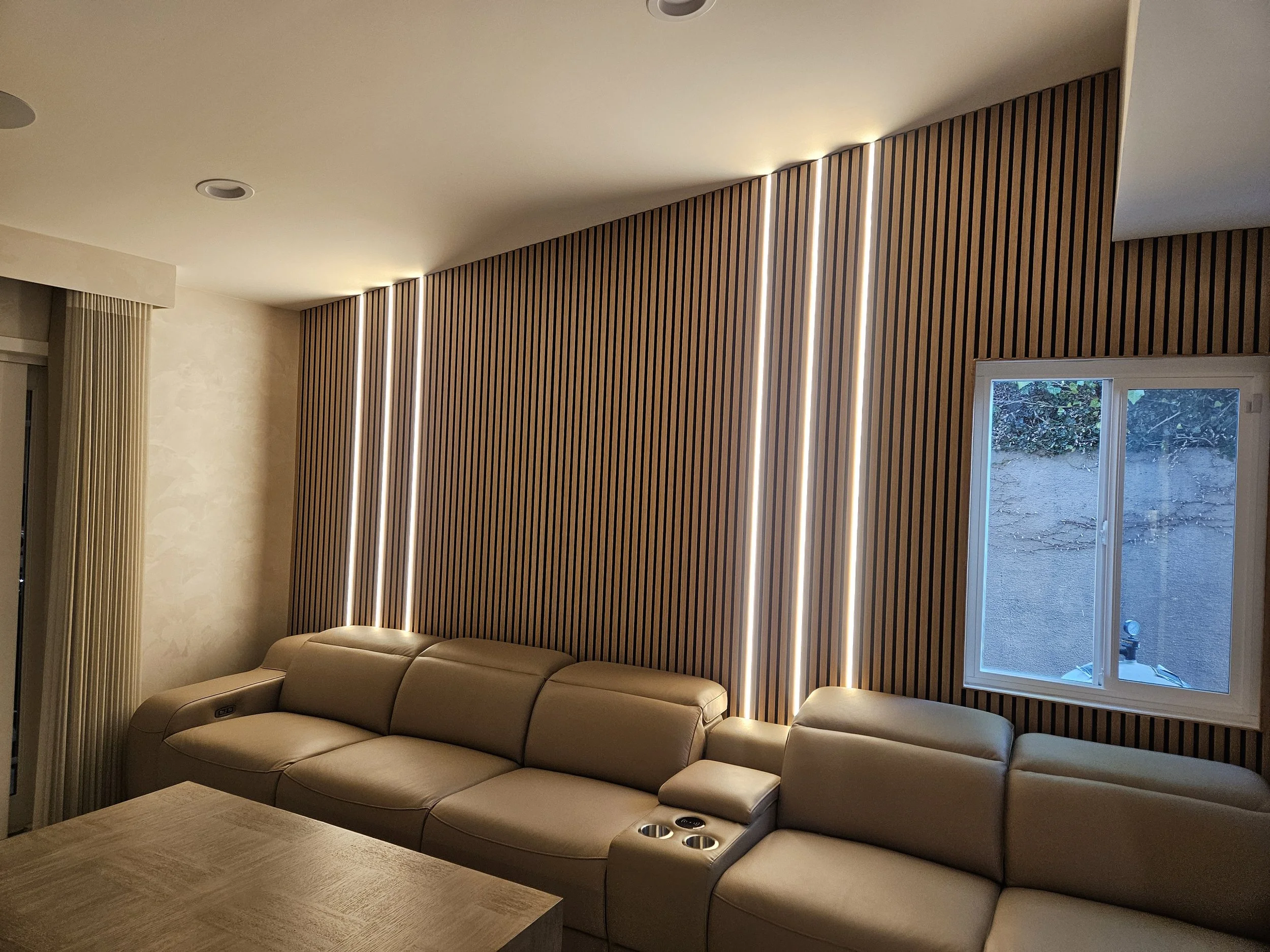 A modern living room with a curved beige leather sectional sofa, a wooden table, a textured beige wall, and a wooden slat accent wall with built-in vertical LED light strips. A window shows an outdoor scene with trees and a garden.